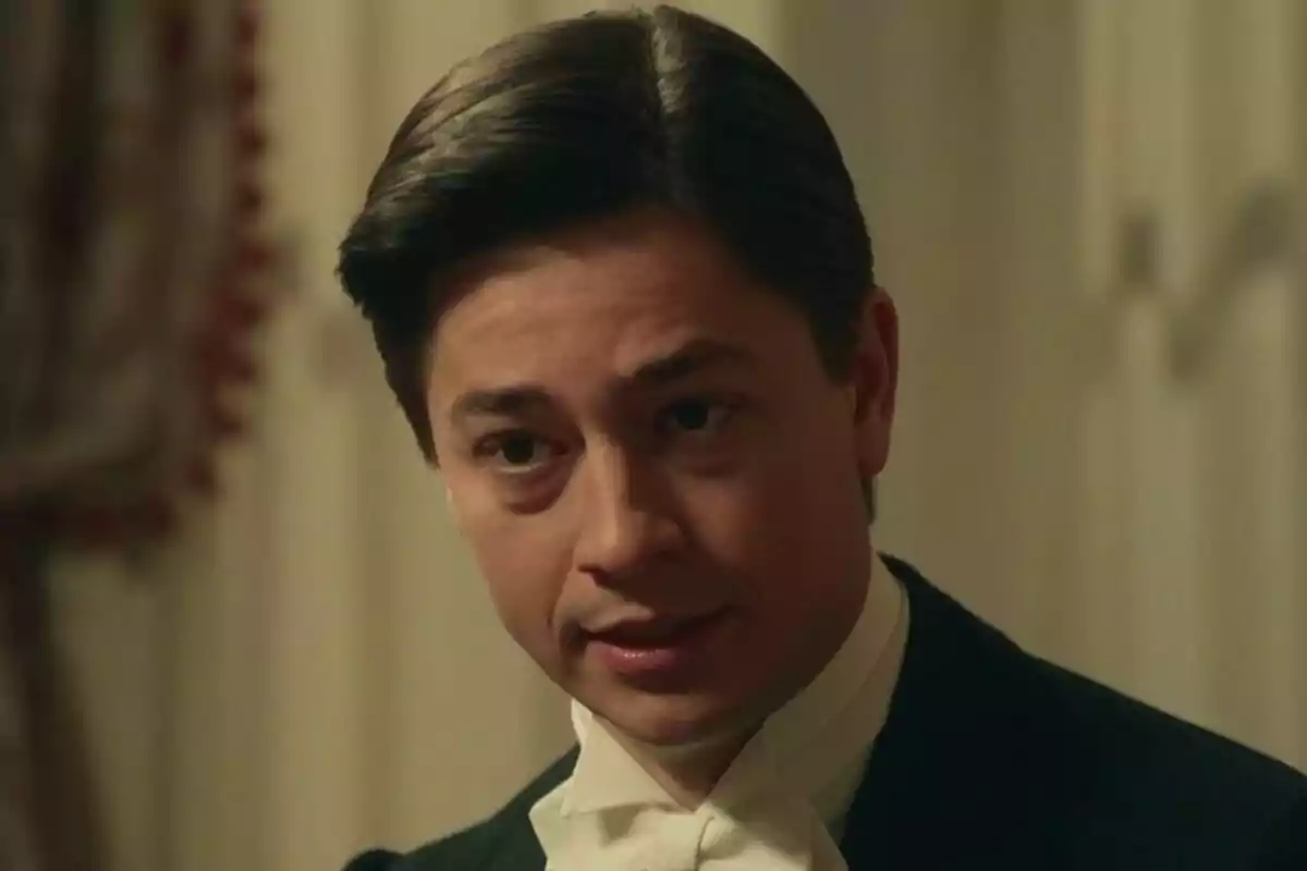 Michel Tejerina con cabello oscuro y peinado hacia atrás, vistiendo un traje formal con corbata blanca, en un ambiente interior en La Promesa. Michel Tejerina con cabello oscuro y peinado hacia atrás, vistiendo un traje formal con corbata blanca, en un ambiente interior en La Promesa.