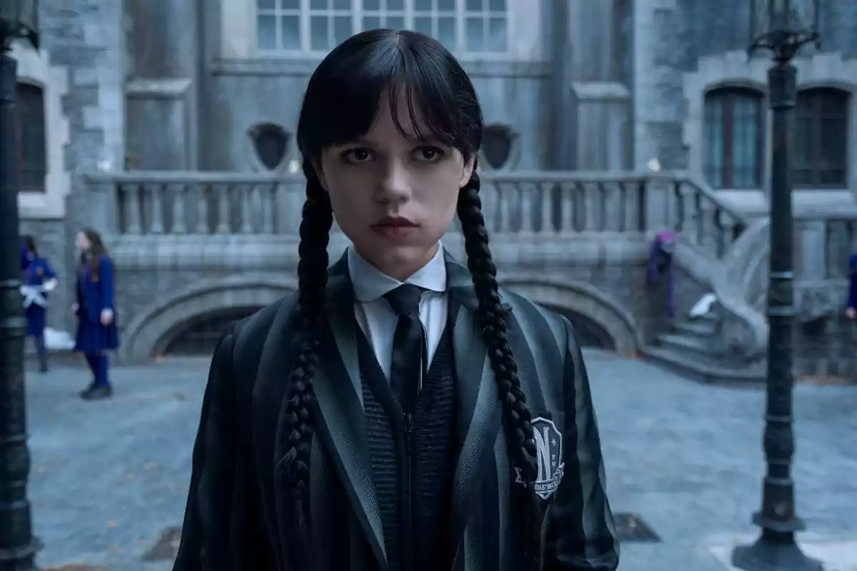 Jenna Ortega con trenzas y expresión seria está de pie frente a un edificio de estilo gótico, vistiendo un uniforme oscuro con corbata en Miércoles.