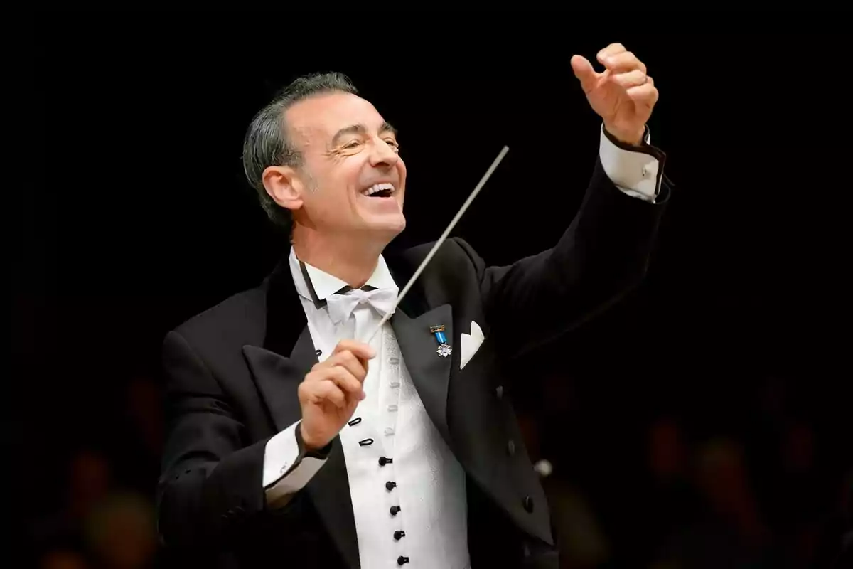 Fotografía de Miguel Ángel Gómez-Martínez como director de orquesta sonriente dirigiendo con una batuta en la mano. Fotografía de Miguel Ángel Gómez-Martínez como director de orquesta sonriente dirigiendo con una batuta en la mano.
