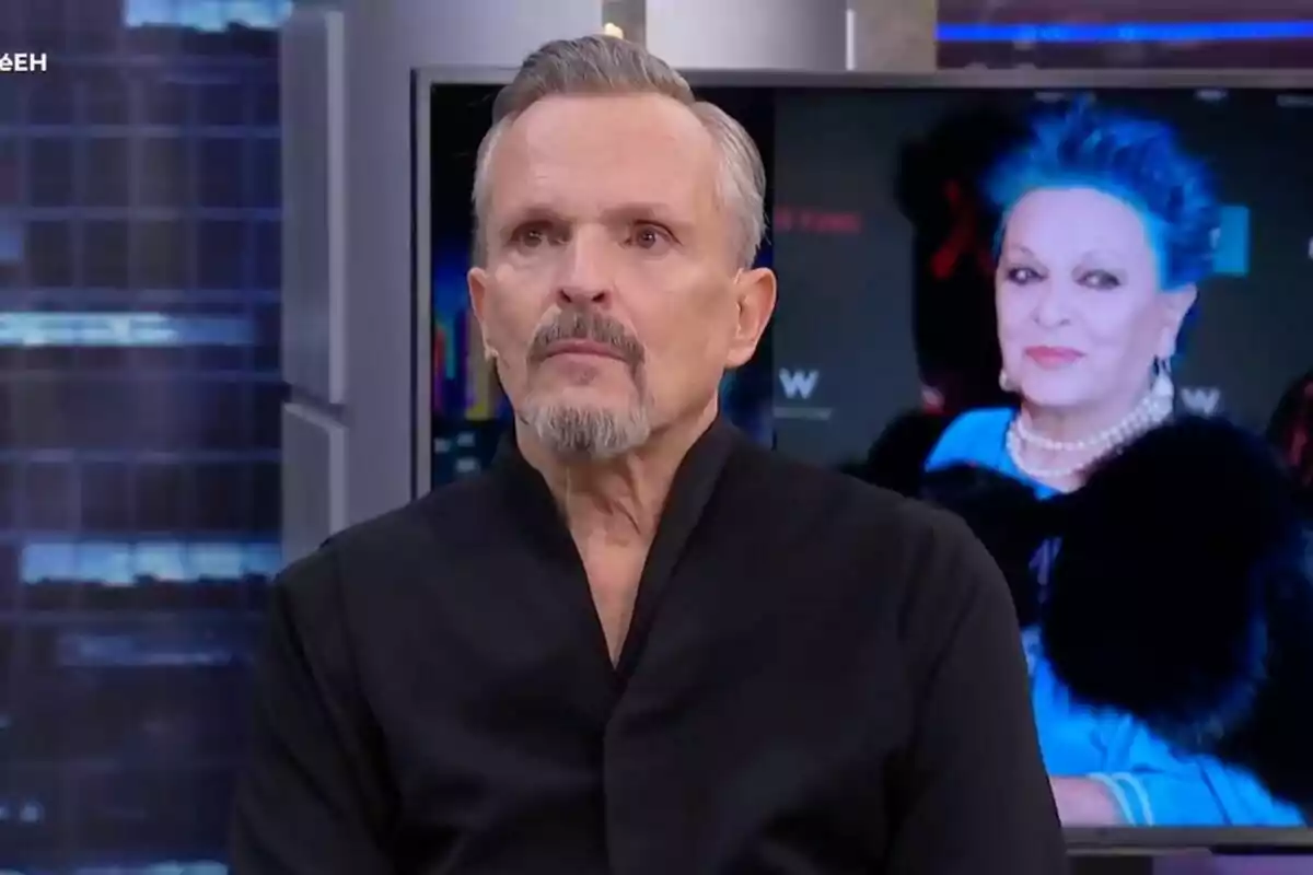 Miguel Bosé con barba y cabello corto está en 'El Hormiguero', detrás de él hay una pantalla que muestra a su madre con cabello azul y un collar de perlas.