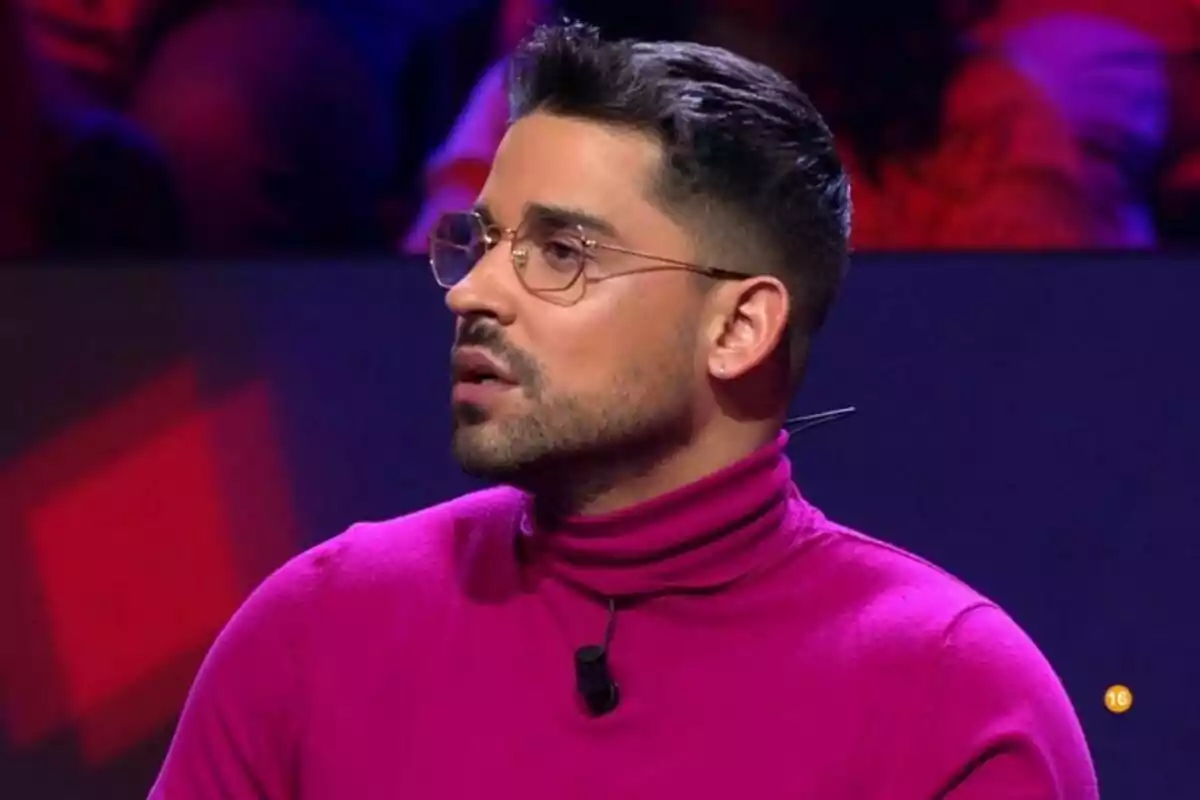 Miguel Frigenti amb ulleres i barba porta un suèter fúcsia i està en un entorn il·luminat amb llums de colors a Gran Hermano. Miguel Frigenti amb ulleres i barba porta un suèter fúcsia i està en un entorn il·luminat amb llums de colors a Gran Hermano.