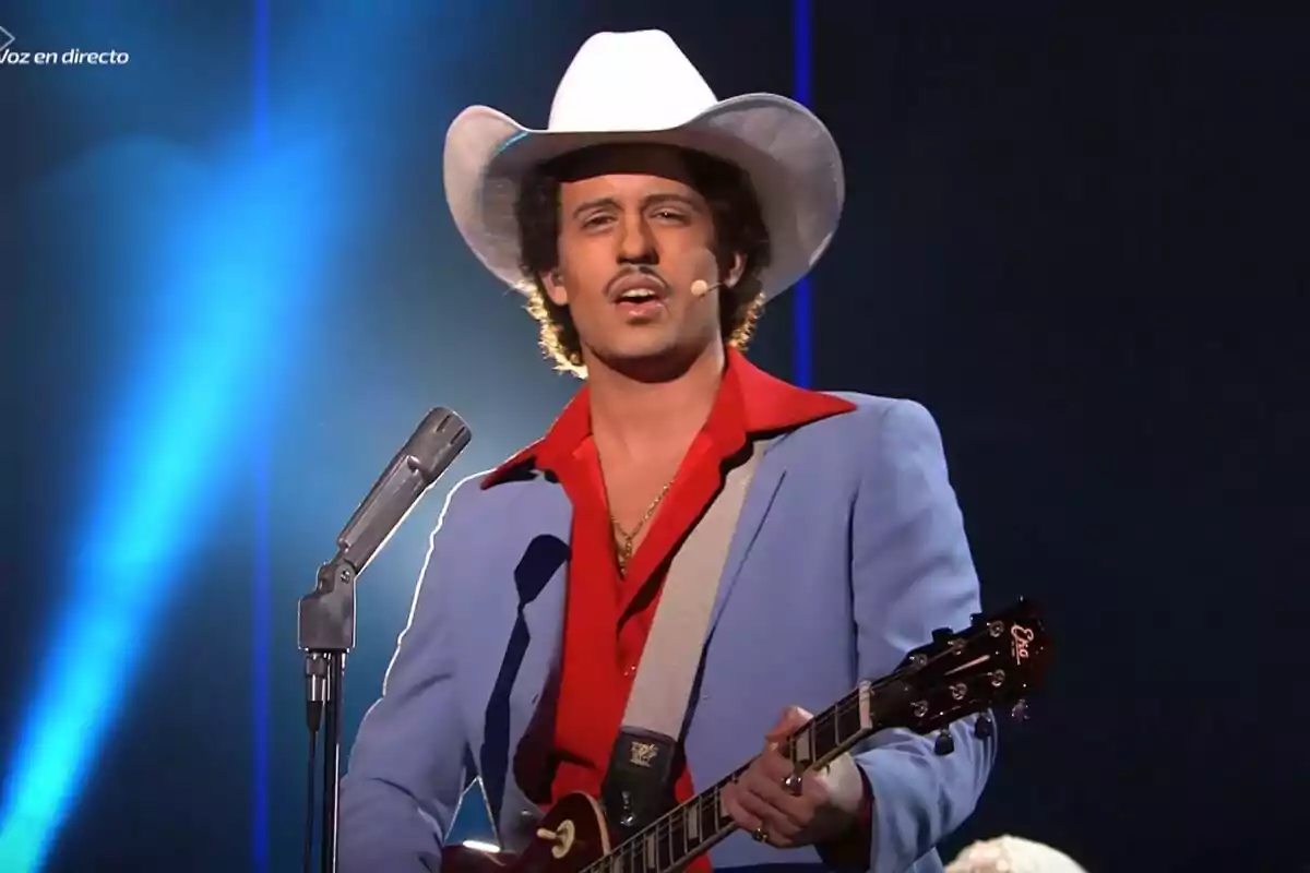 Mikel Herzog JR con sombrero blanco y traje azul claro toca la guitarra en un escenario iluminado en Tu Cara Me Suena.