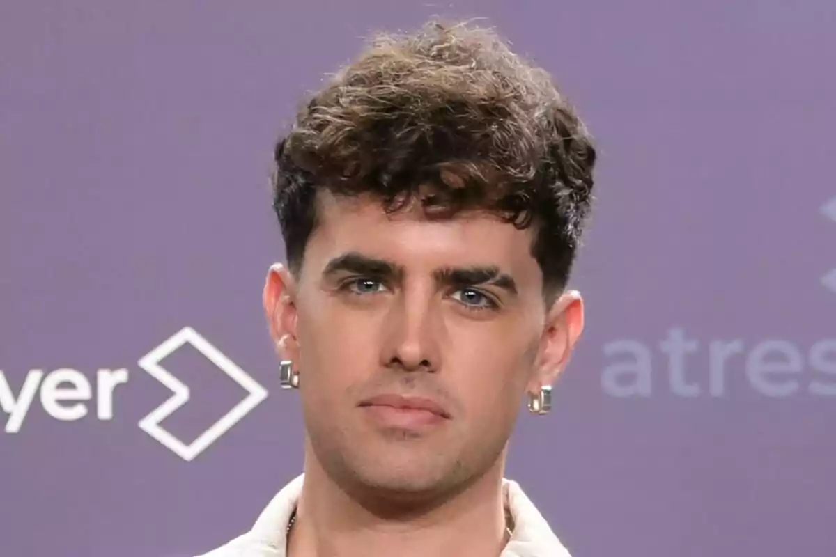 Mikel Herzog JR con cabello rizado y aretes posando frente a un fondo morado con logotipos de Tu Cara Me Suena.