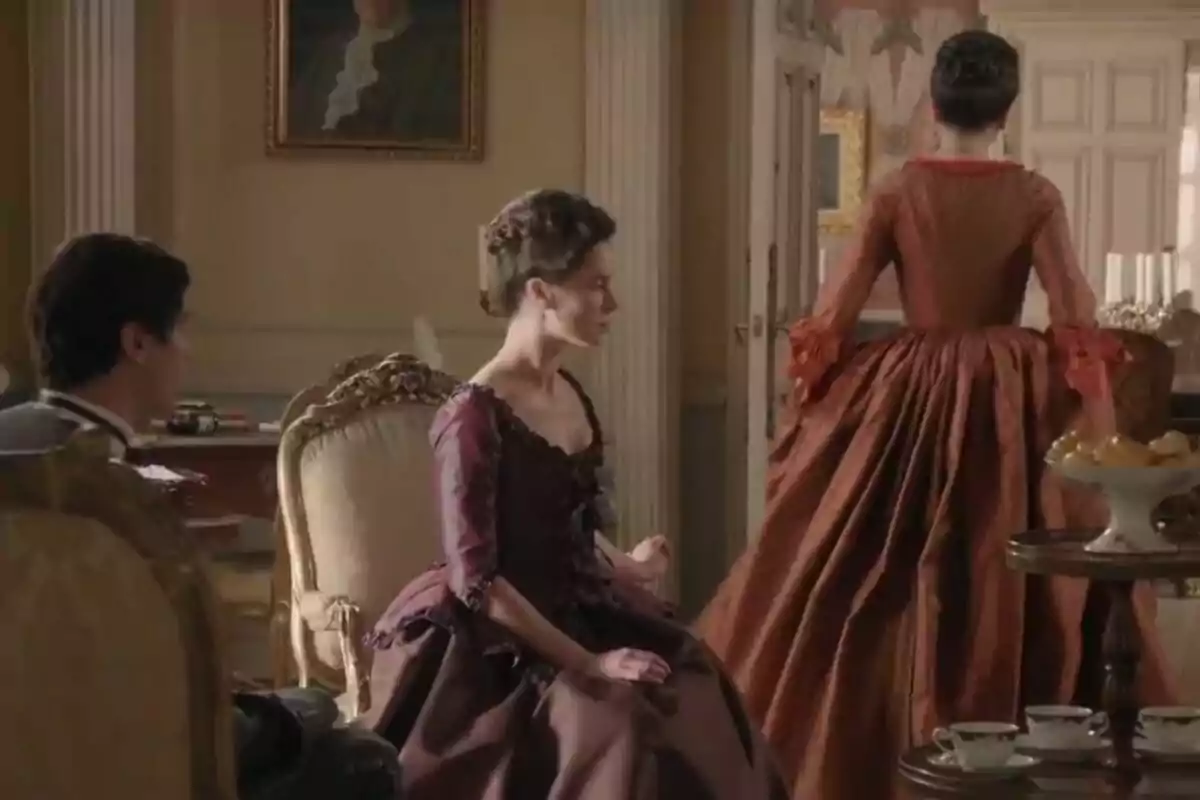 Tres personas en una sala elegante con vestimenta de época, una de ellas de pie y las otras dos sentadas, rodeadas de muebles antiguos y una mesa con tazas de té. Tres personas en una sala elegante con vestimenta de época, una de ellas de pie y las otras dos sentadas, rodeadas de muebles antiguos y una mesa con tazas de té.