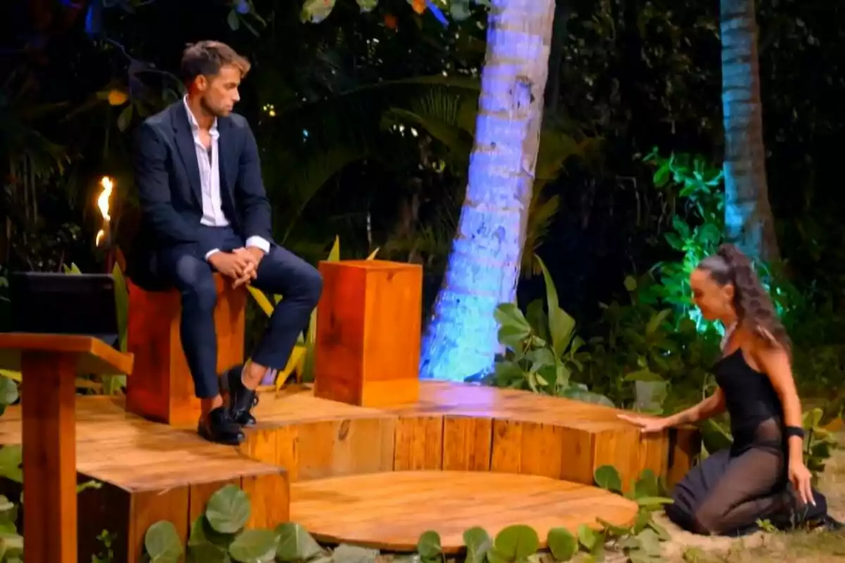 Montoya con traje está sentado en un escenario de madera al aire libre mientras observa a Anita Williams que se arrodilla en el suelo, rodeados de vegetación y luces de colores en La Isla de las Tentaciones. Montoya con traje está sentado en un escenario de madera al aire libre mientras observa a Anita Williams que se arrodilla en el suelo, rodeados de vegetación y luces de colores en La Isla de las Tentaciones.