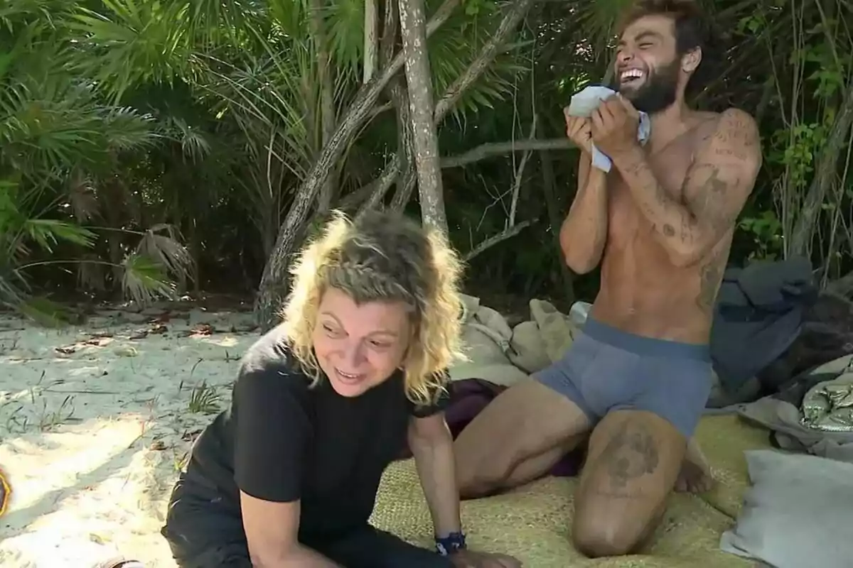 Dos personas en una playa de Supervivientes, Terelu Campos sentada en la arena y Montoya de pie riendo, rodeadas de vegetación. Dos personas en una playa de Supervivientes, Terelu Campos sentada en la arena y Montoya de pie riendo, rodeadas de vegetación.