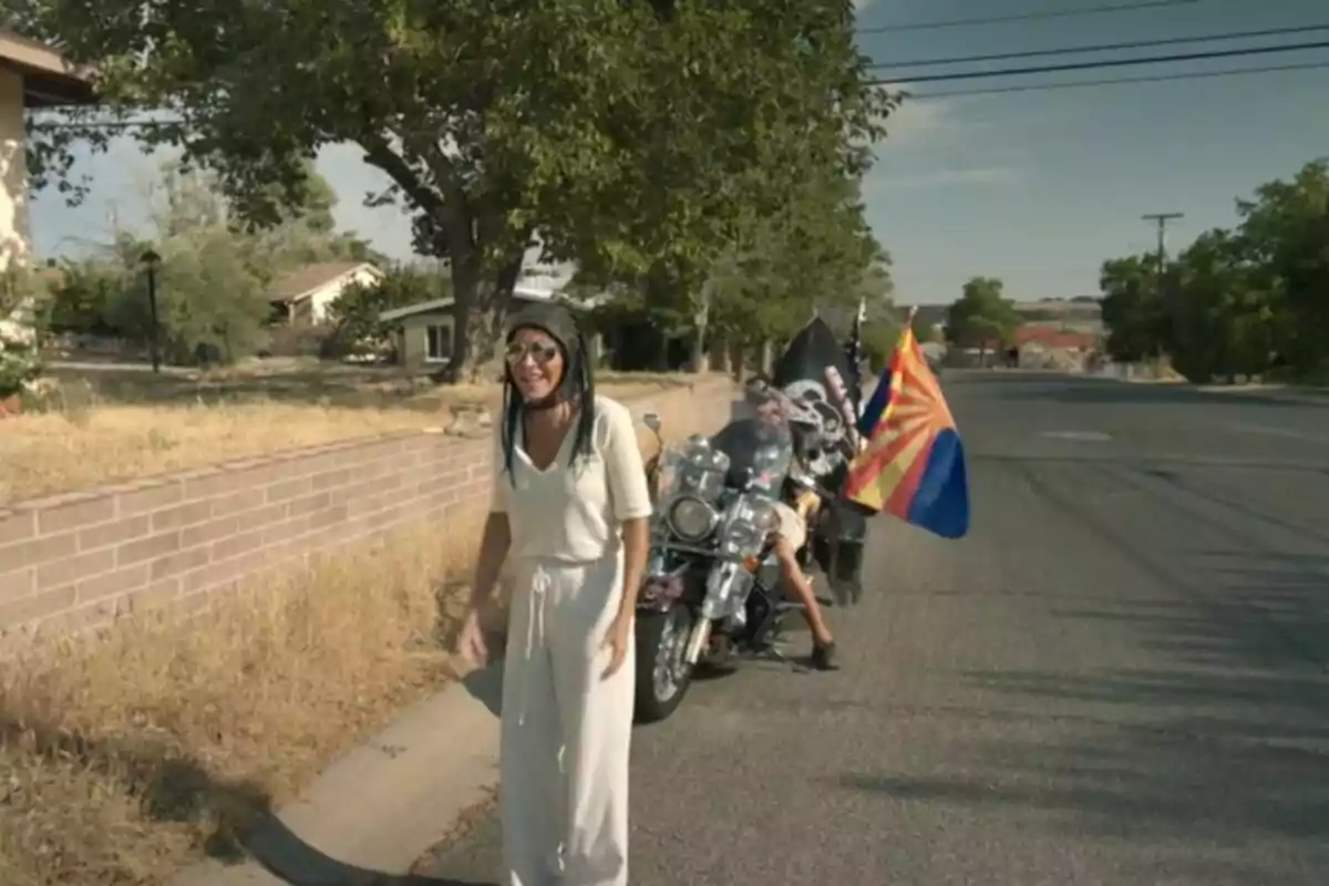 Susana Saborido sonriente con casco y gafas de sol camina por una calle mientras dos motociclistas con banderas están estacionados detrás. Susana Saborido sonriente con casco y gafas de sol camina por una calle mientras dos motociclistas con banderas están estacionados detrás.