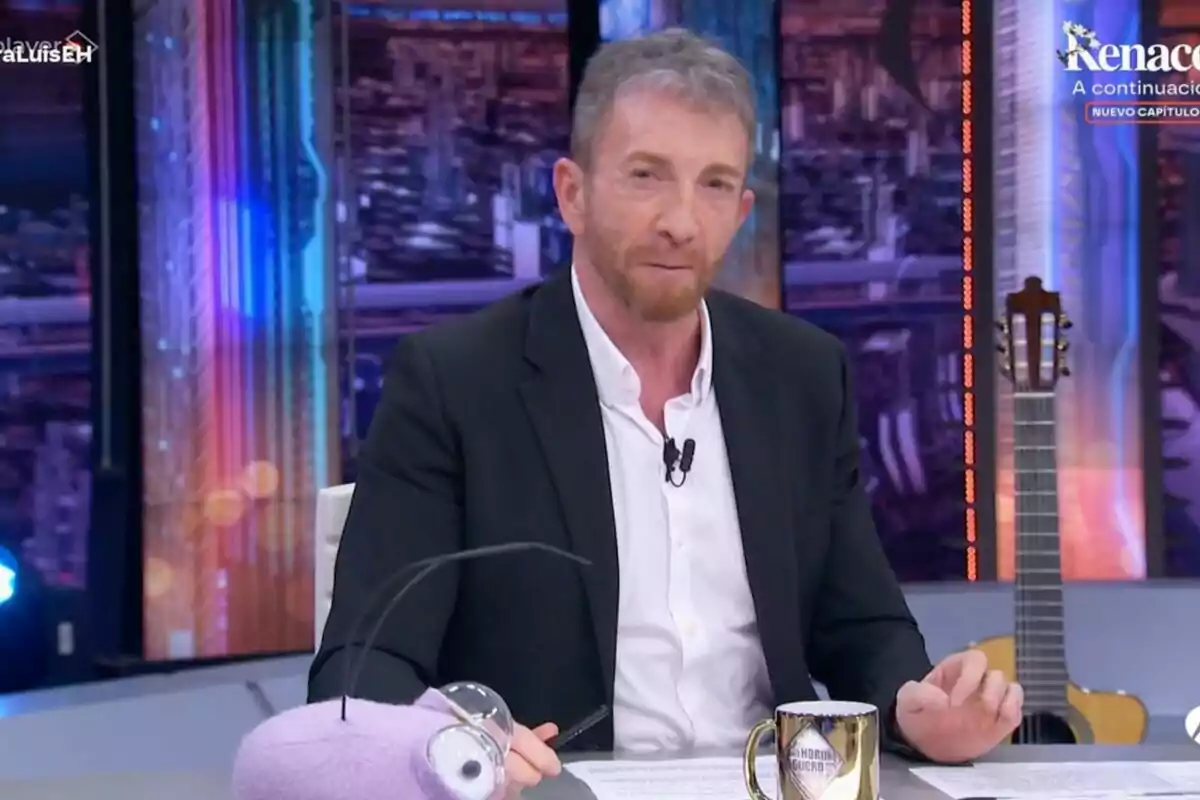 Pablo Motos amb barba i vestit fosc està assegut a 'El Hormiguero', davant seu hi ha una tassa i un peluix morat amb antenes, al fons es veu una guitarra i llums de colors. Pablo Motos amb barba i vestit fosc està assegut a 'El Hormiguero', davant seu hi ha una tassa i un peluix morat amb antenes, al fons es veu una guitarra i llums de colors.