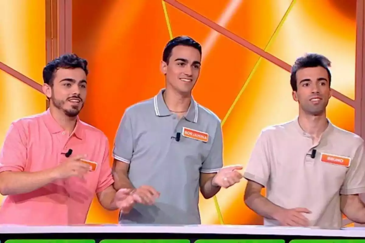 Los Mozos de Arousa de pie en un set de televisión con fondo naranja, cada uno con una camiseta de diferente color y una etiqueta con su nombre. Los Mozos de Arousa de pie en un set de televisión con fondo naranja, cada uno con una camiseta de diferente color y una etiqueta con su nombre.