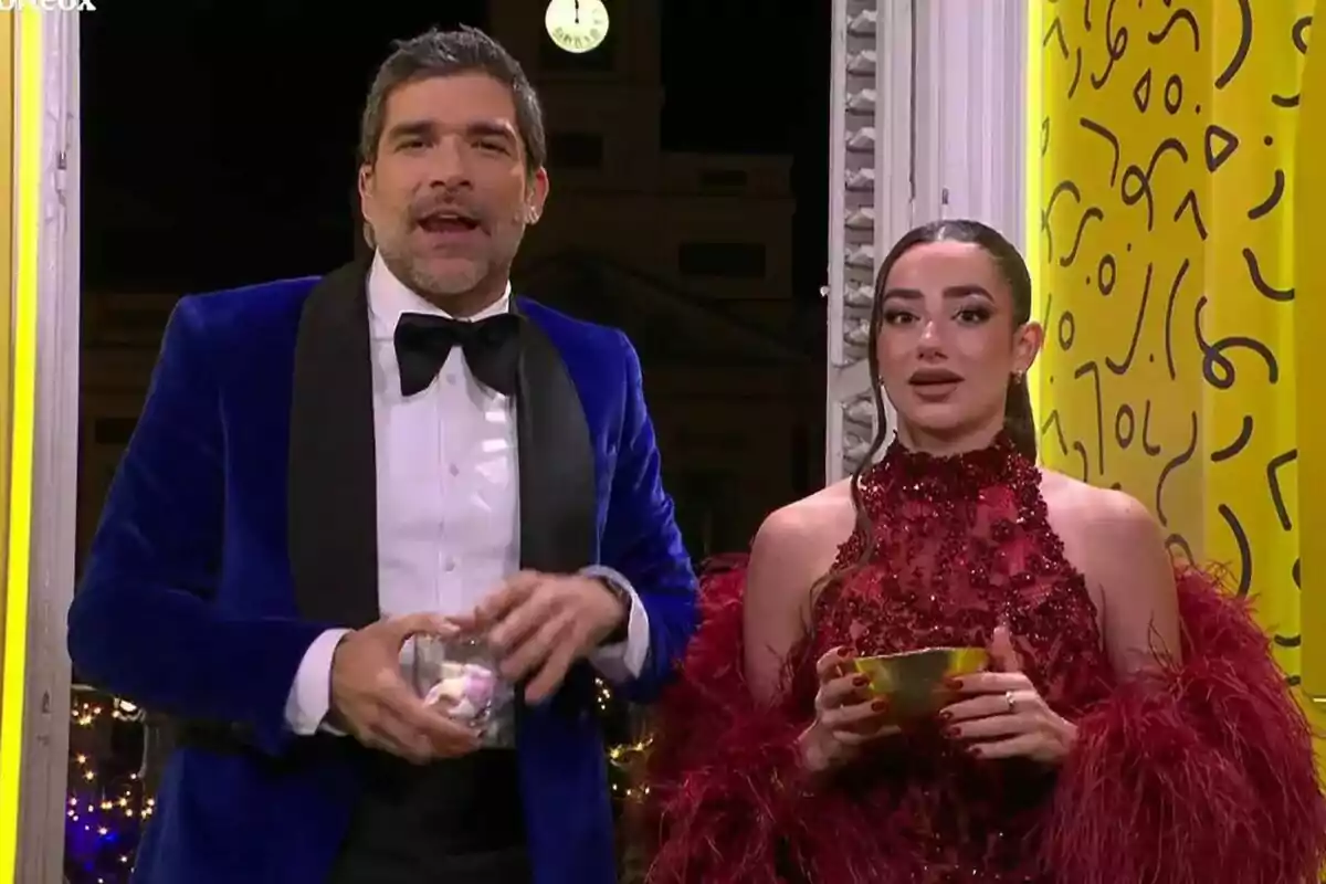 Dues persones elegantment vestides en un esdeveniment nocturn, una amb un esmòquing blau i l'altra amb un vestit vermell brillant, sostenint recipients a les mans. Dues persones elegantment vestides en un esdeveniment nocturn, una amb un esmòquing blau i l'altra amb un vestit vermell brillant, sostenint recipients a les mans.