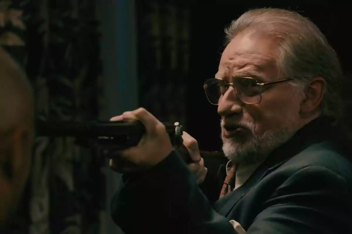 Nancho Novo mayor con barba y gafas sostiene una escopeta apuntando a otra persona en un entorno oscuro en Sueños de Libertad. Nancho Novo mayor con barba y gafas sostiene una escopeta apuntando a otra persona en un entorno oscuro en Sueños de Libertad.