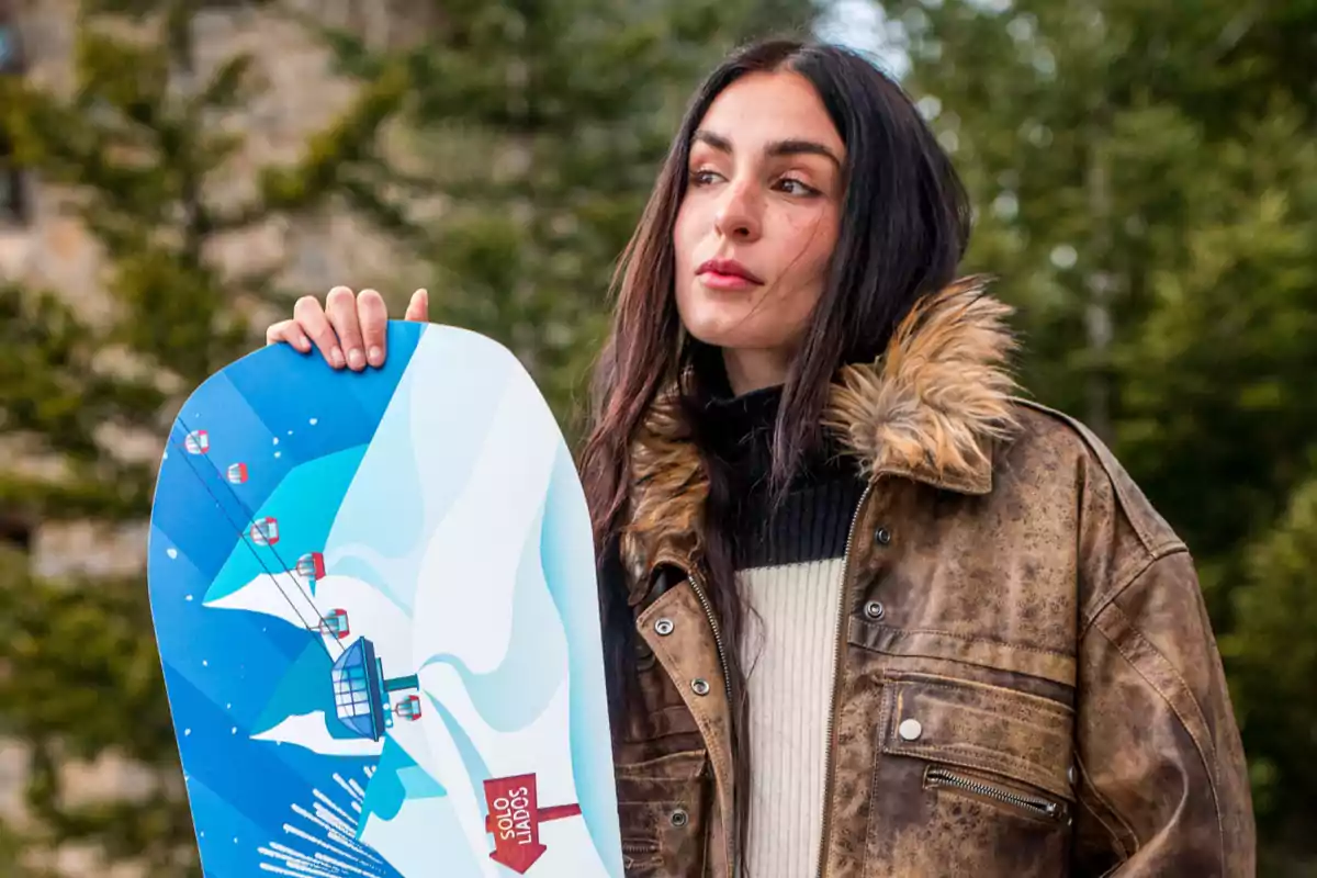 Natalia Lacunza amb jaqueta de cuir i caputxa de pell sosté una taula de snowboard amb un disseny de telefèric en un entorn natural. Natalia Lacunza amb jaqueta de cuir i caputxa de pell sosté una taula de snowboard amb un disseny de telefèric en un entorn natural.