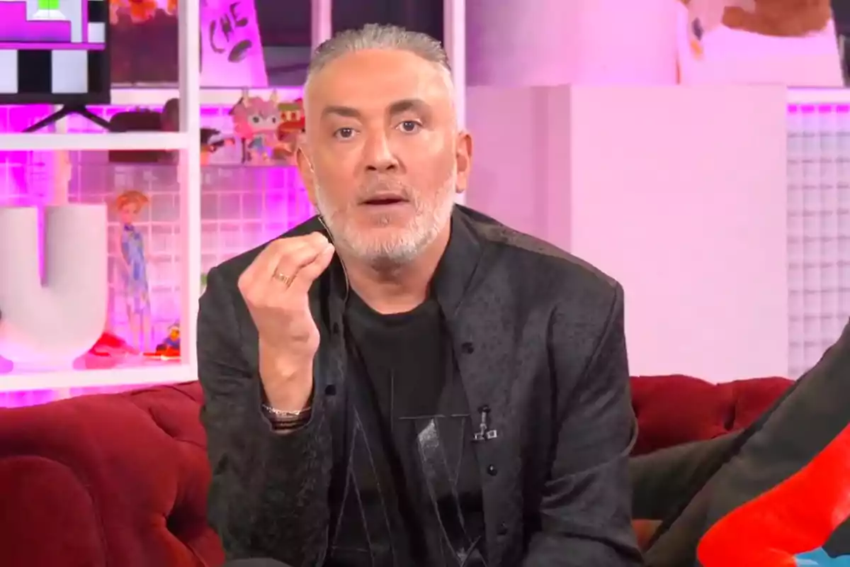 Kiko Hernández con barba y cabello canoso está sentado en un sofá rojo, vistiendo una chaqueta oscura, con un fondo iluminado en tonos rosados y objetos decorativos. Captura de 'Ni que fuéramos' Kiko Hernández con barba y cabello canoso está sentado en un sofá rojo, vistiendo una chaqueta oscura, con un fondo iluminado en tonos rosados y objetos decorativos. Captura de 'Ni que fuéramos'