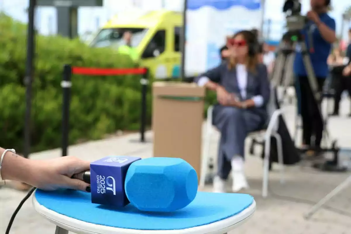 Una mano sostiene un micrófono azul con un logotipo mientras en el fondo se ve a Noemí Galera sentada y una cámara de video en un entorno al aire libre en el casting de OT 2025.