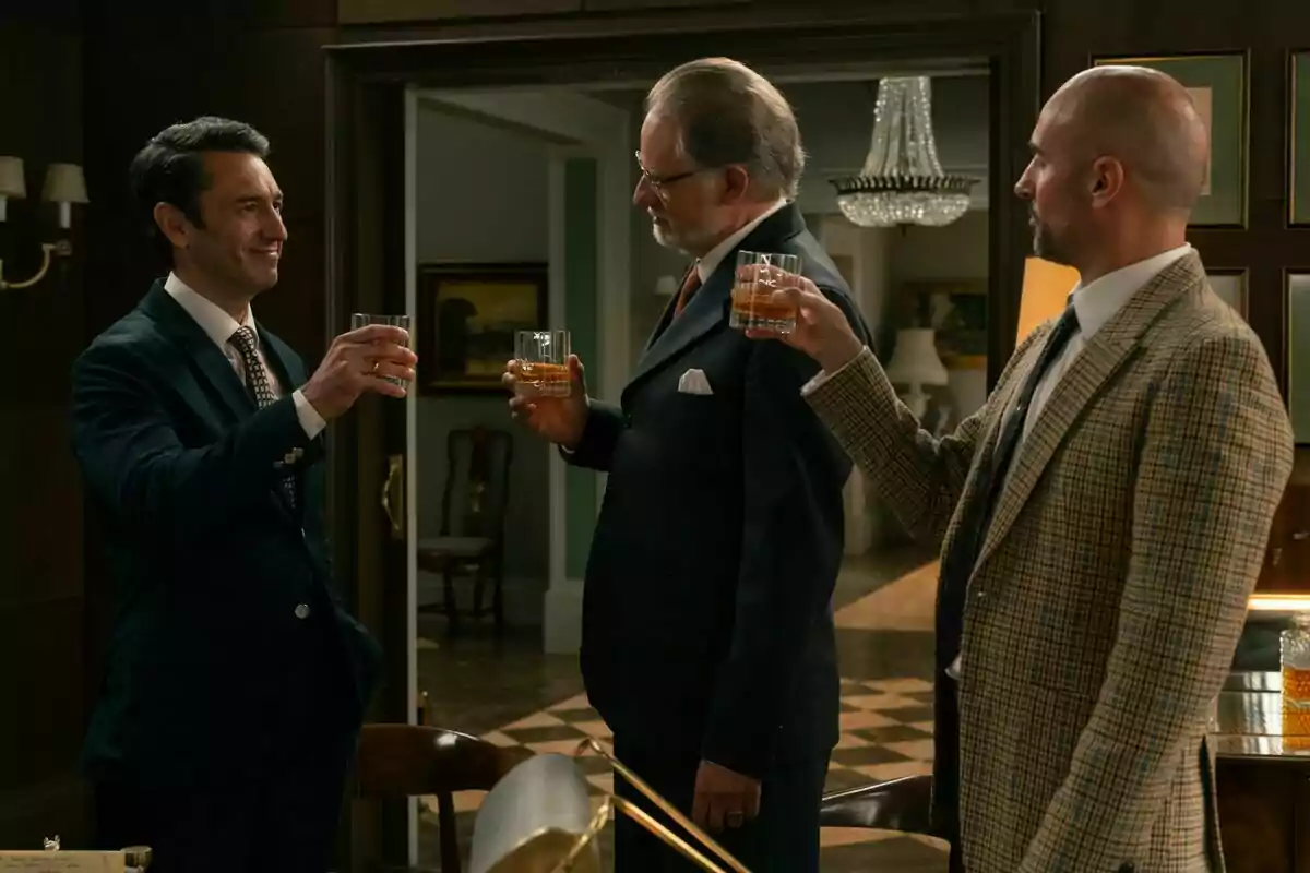 Ricardo Reguera, Nancho Novo y Alain Hernández bien vestidos brindan con vasos de whisky en una elegante sala con una lámpara de araña al fondo en Sueños de Libertad. Ricardo Reguera, Nancho Novo y Alain Hernández bien vestidos brindan con vasos de whisky en una elegante sala con una lámpara de araña al fondo en Sueños de Libertad.