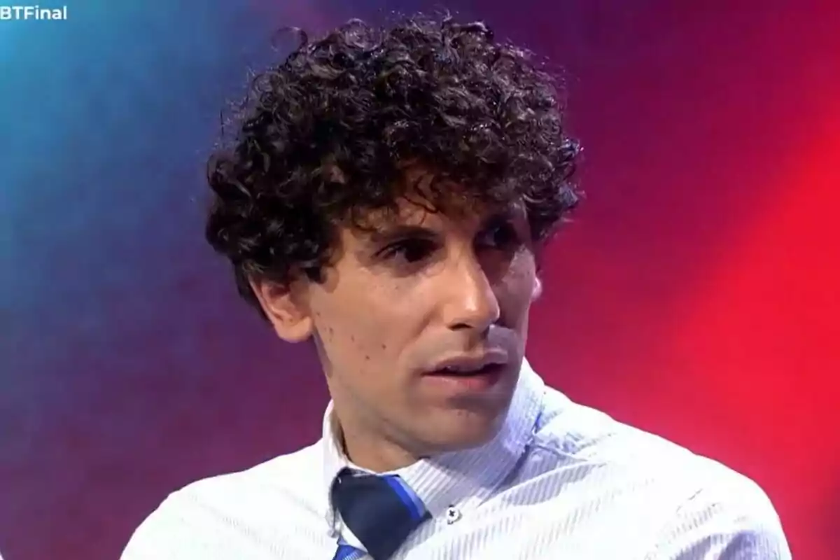 Óscar Landa con cabello rizado y expresión seria viste camisa blanca y corbata azul sobre un fondo de colores rojo y azul en Gran Hermano. Óscar Landa con cabello rizado y expresión seria viste camisa blanca y corbata azul sobre un fondo de colores rojo y azul en Gran Hermano.