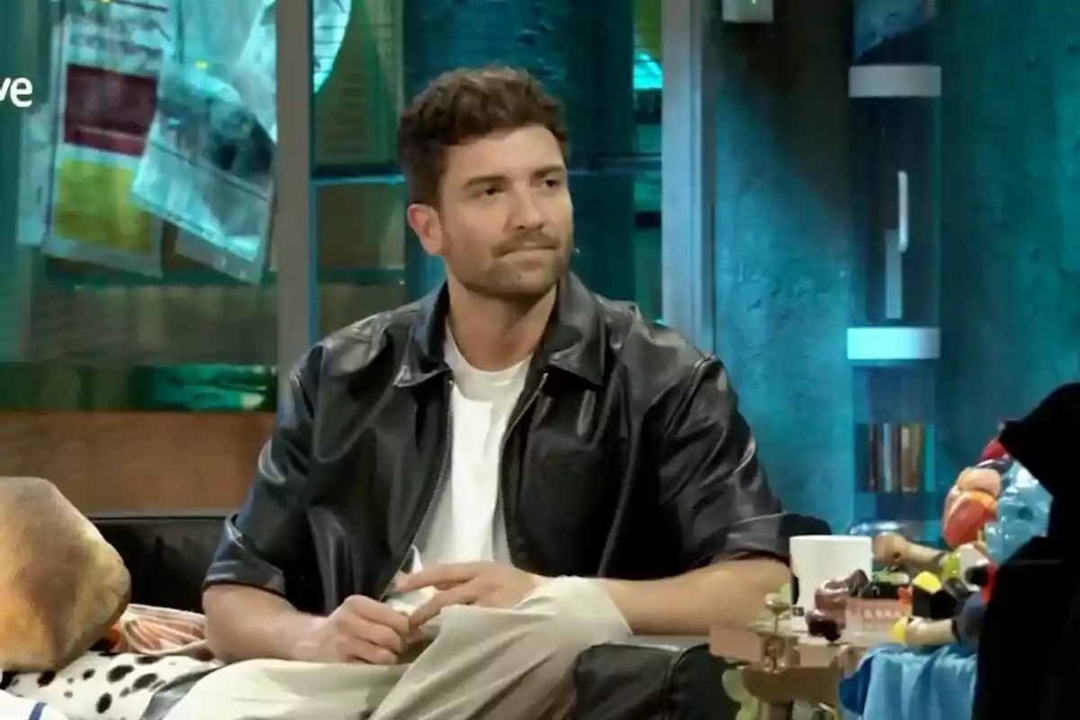Pablo Alborán con chaqueta de cuero negra y camiseta blanca está sentado en un sofá en un ambiente moderno y colorido en 'La Revuelta'.