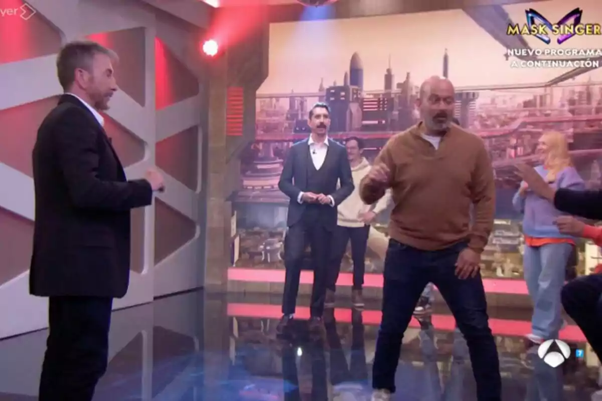 Un grupo de personas como Pablo Motos en El Hormiguero con luces rojas y una pantalla de fondo que muestra una ciudad futurista. Un grupo de personas como Pablo Motos en El Hormiguero con luces rojas y una pantalla de fondo que muestra una ciudad futurista.