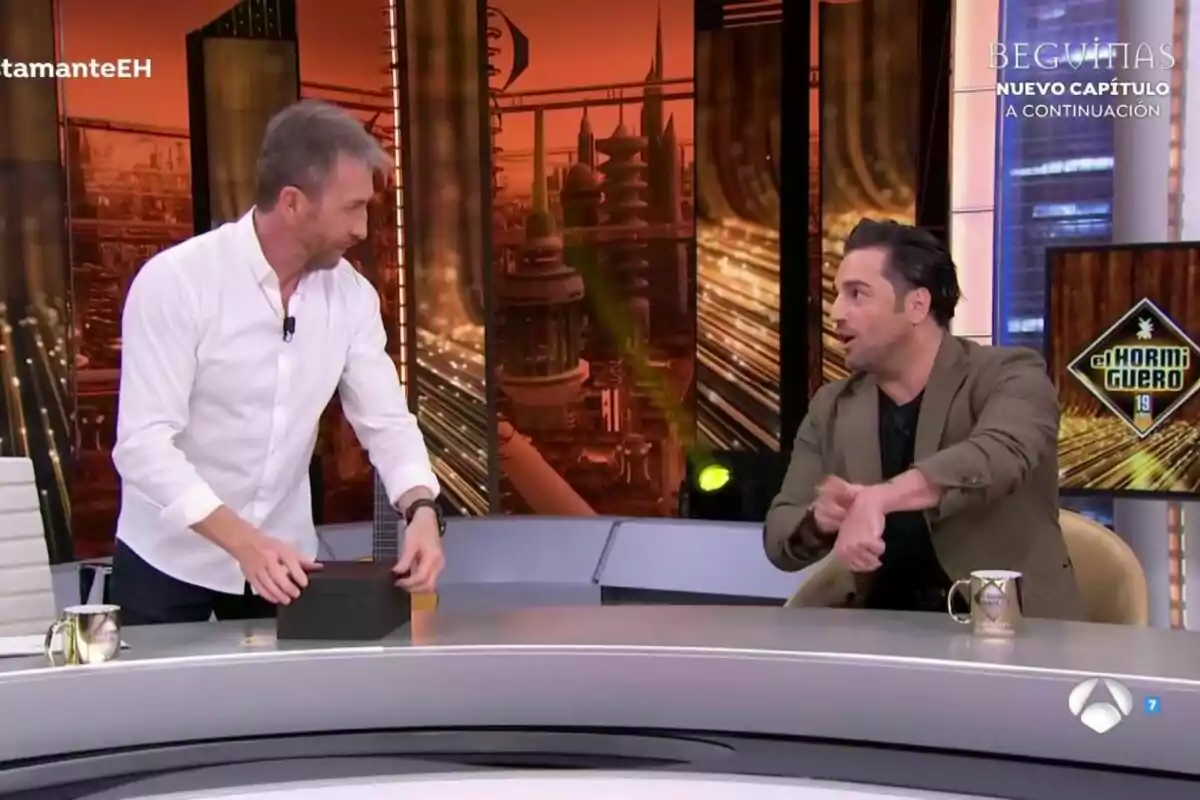 Pablo Motos y David Bustamante conversan animadamente en El Hormiguero con un fondo futurista y una pantalla que muestra el logo de un programa. Pablo Motos y David Bustamante conversan animadamente en El Hormiguero con un fondo futurista y una pantalla que muestra el logo de un programa.