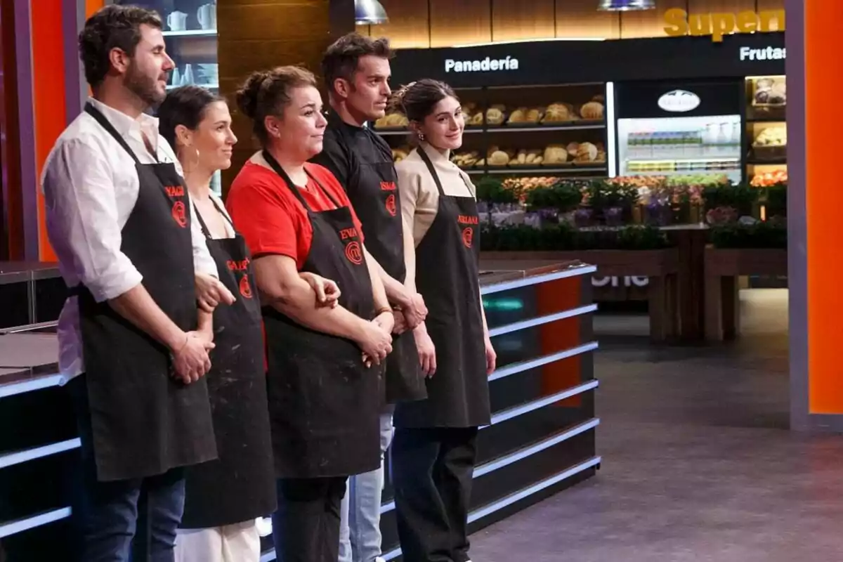 Cinco personas con delantales negros están de pie en una cocina de 'Masterchef', con una panadería visible al fondo.