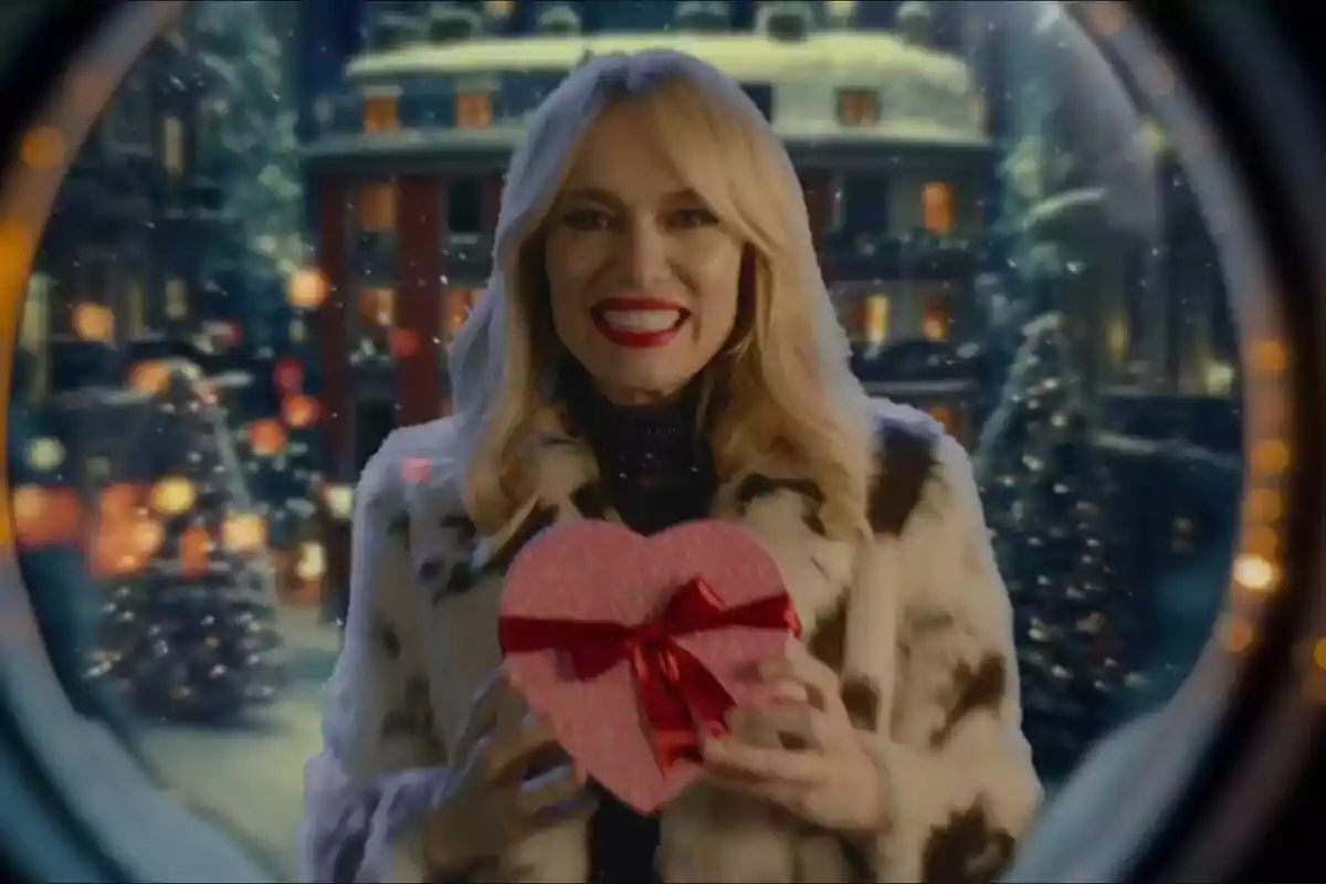 Patricia Conde sonriente con un abrigo de piel sostiene una caja de regalo en forma de corazón con un lazo rojo, en un entorno invernal con luces y nieve. Patricia Conde sonriente con un abrigo de piel sostiene una caja de regalo en forma de corazón con un lazo rojo, en un entorno invernal con luces y nieve.