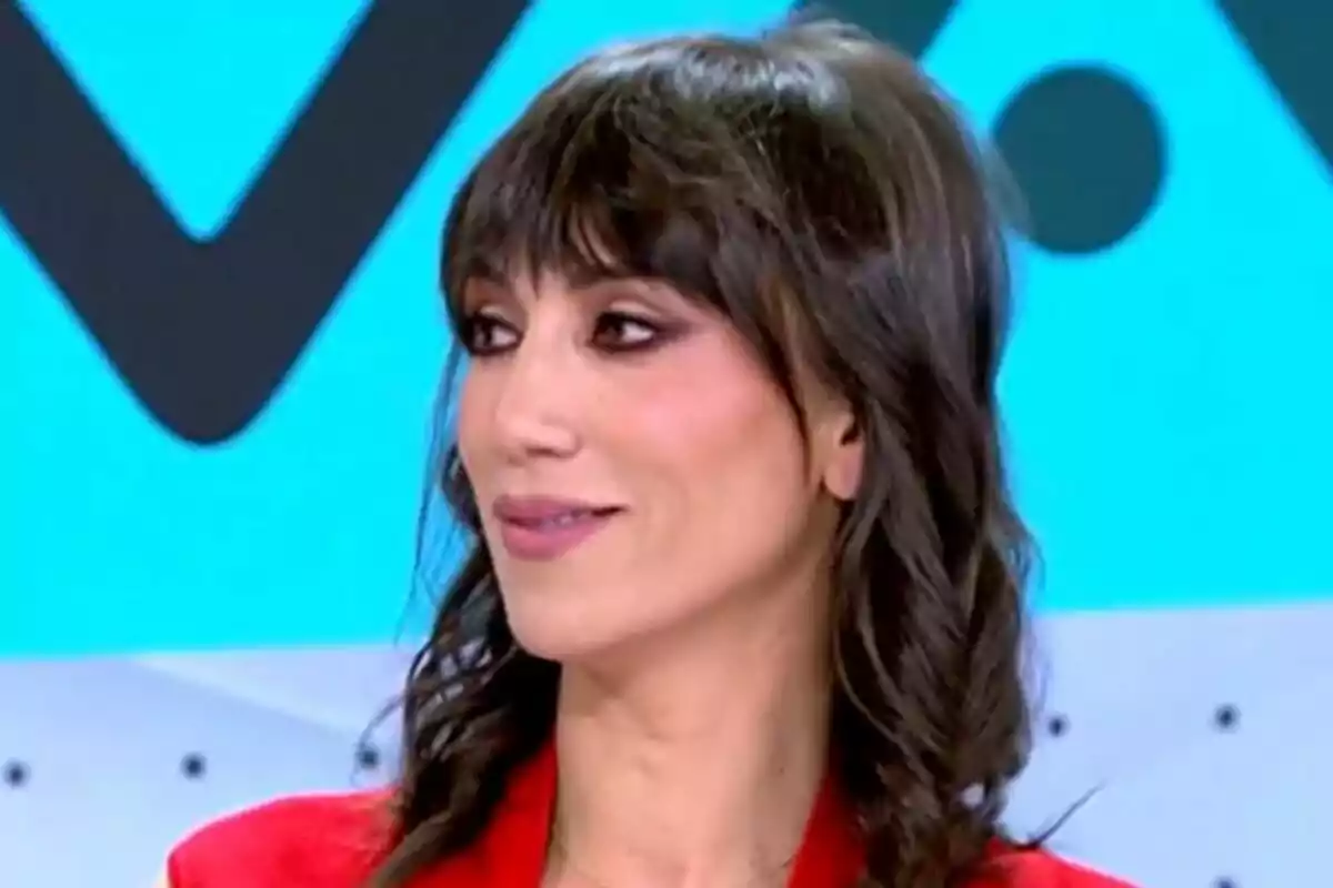 Patricia Pardo con cabello oscuro y rizado, vistiendo una chaqueta roja, está sonriendo y mirando hacia un lado, con un fondo azul claro y formas geométricas en Vamos a ver.