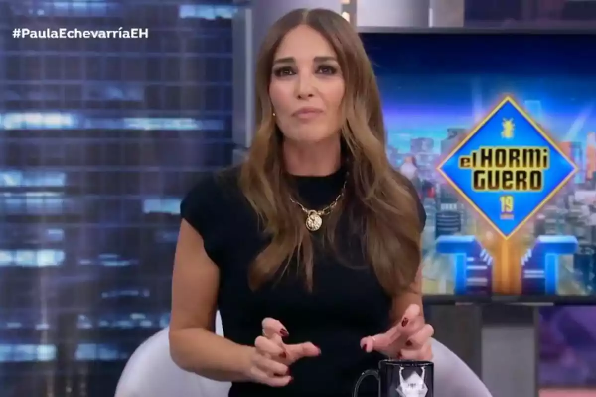 Paula Echevarría con cabello largo y castaño está sentada en un set de televisión, vestida de negro y con un collar dorado, mientras sostiene una taza negra. Al fondo, se ve el logotipo del programa Paula Echevarría con cabello largo y castaño está sentada en un set de televisión, vestida de negro y con un collar dorado, mientras sostiene una taza negra. Al fondo, se ve el logotipo del programa