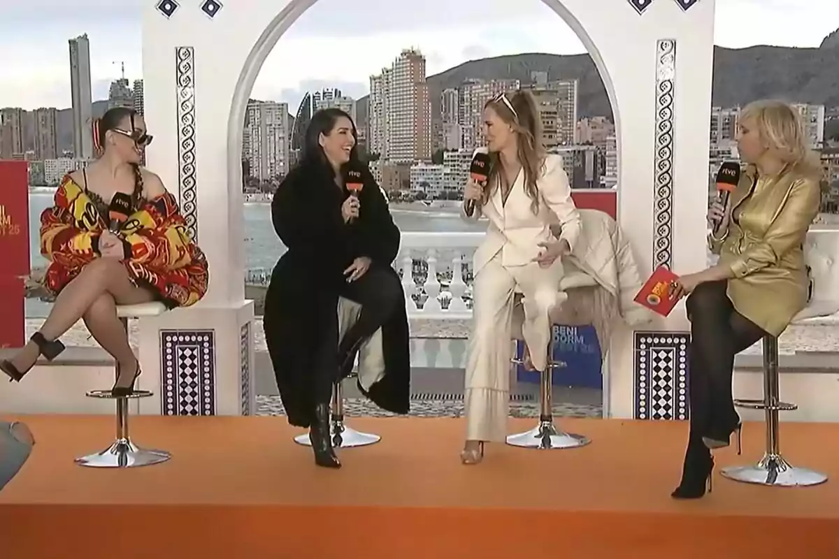 Inés Hernand, Ruth Lorenzo, Paula Vázquez y María Eizaguirre sentadas en taburetes altos en la presentación del Benidorm Fest al aire libre con un fondo de edificios y montañas, sosteniendo micrófonos y conversando animadamente. Inés Hernand, Ruth Lorenzo, Paula Vázquez y María Eizaguirre sentadas en taburetes altos en la presentación del Benidorm Fest al aire libre con un fondo de edificios y montañas, sosteniendo micrófonos y conversando animadamente.
