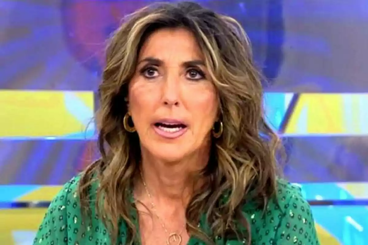 Mujer con cabello ondulado y castaño claro, usando una blusa verde con estampado y aretes dorados, hablando frente a un fondo colorido. Mujer con cabello ondulado y castaño claro, usando una blusa verde con estampado y aretes dorados, hablando frente a un fondo colorido.