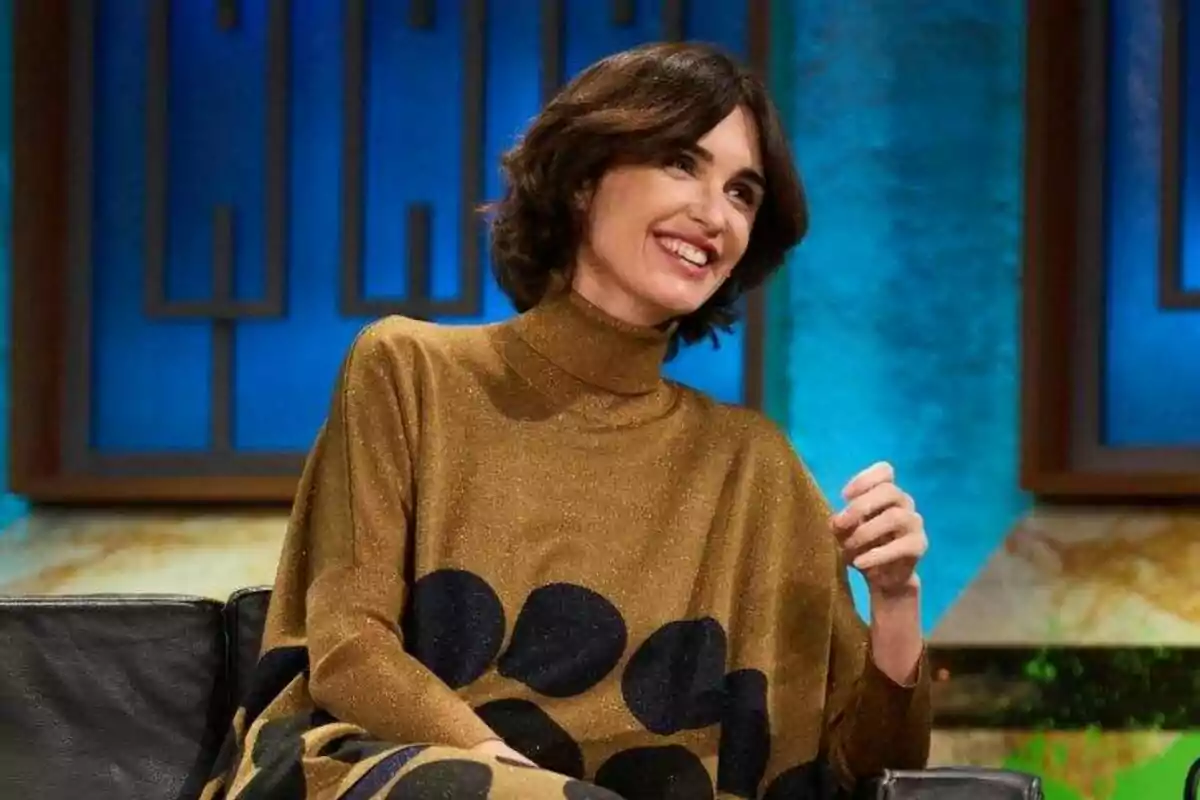 Paz Vega con cabello oscuro y corto sonríe mientras está sentada en un sofá con un fondo azul en La Revuelta. Paz Vega con cabello oscuro y corto sonríe mientras está sentada en un sofá con un fondo azul en La Revuelta.