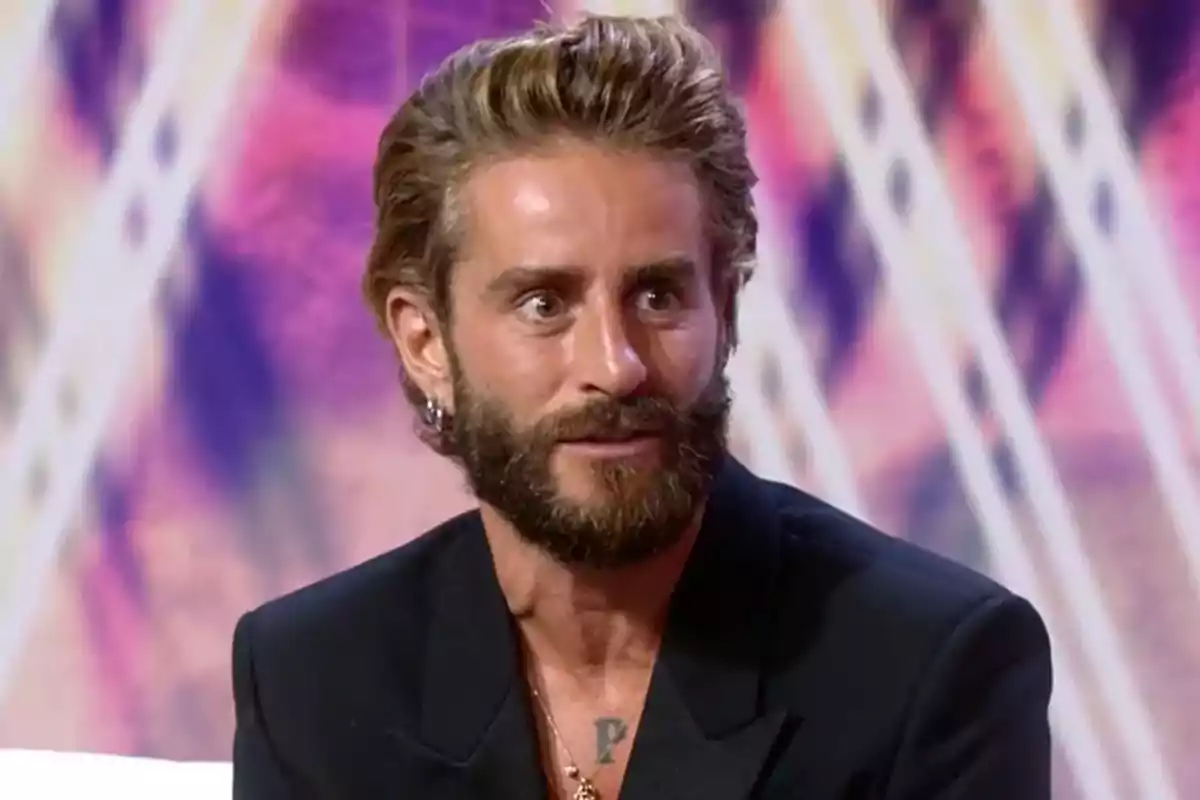 Pelayo Díaz con barba y cabello peinado hacia atrás vistiendo saco oscuro en un set de televisión con fondo colorido