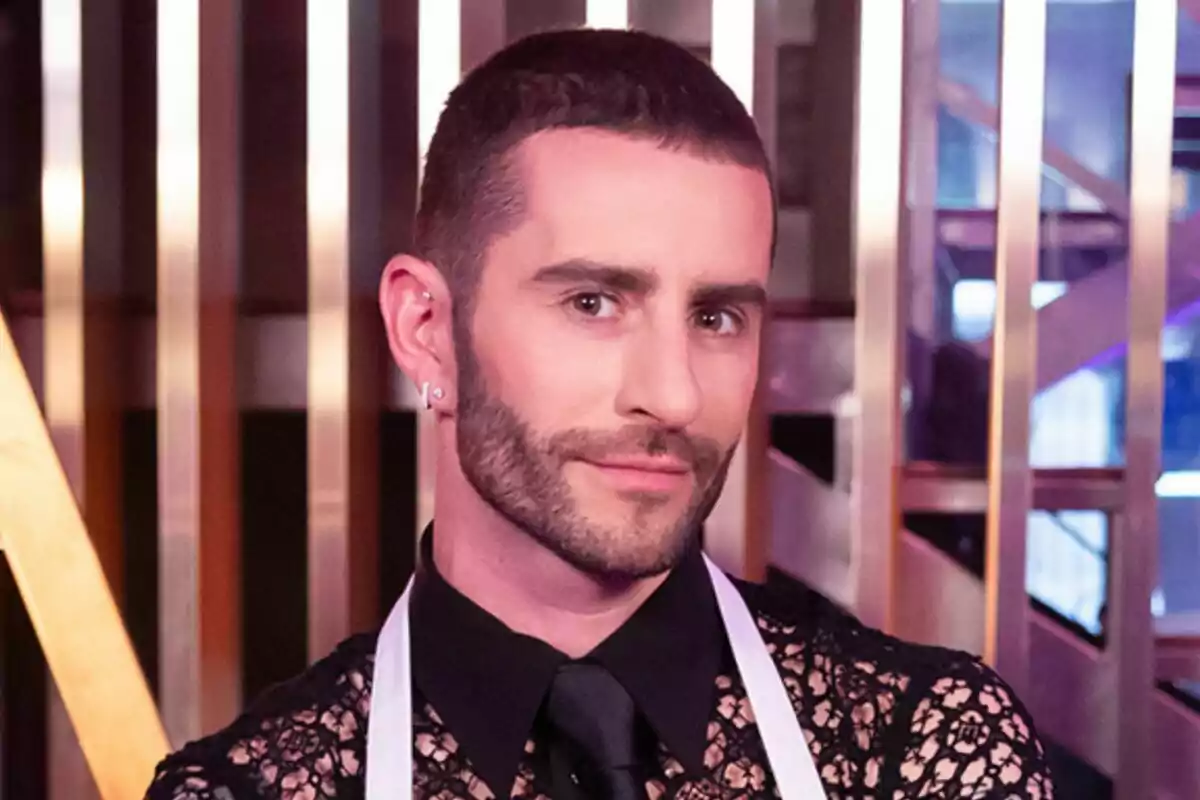 Pelayo Díaz con barba y cabello corto, usando una camisa de encaje negra y una corbata negra, con un delantal blanco, posando frente a una estructura metálica en MasterChef Celebrity. Pelayo Díaz con barba y cabello corto, usando una camisa de encaje negra y una corbata negra, con un delantal blanco, posando frente a una estructura metálica en MasterChef Celebrity.