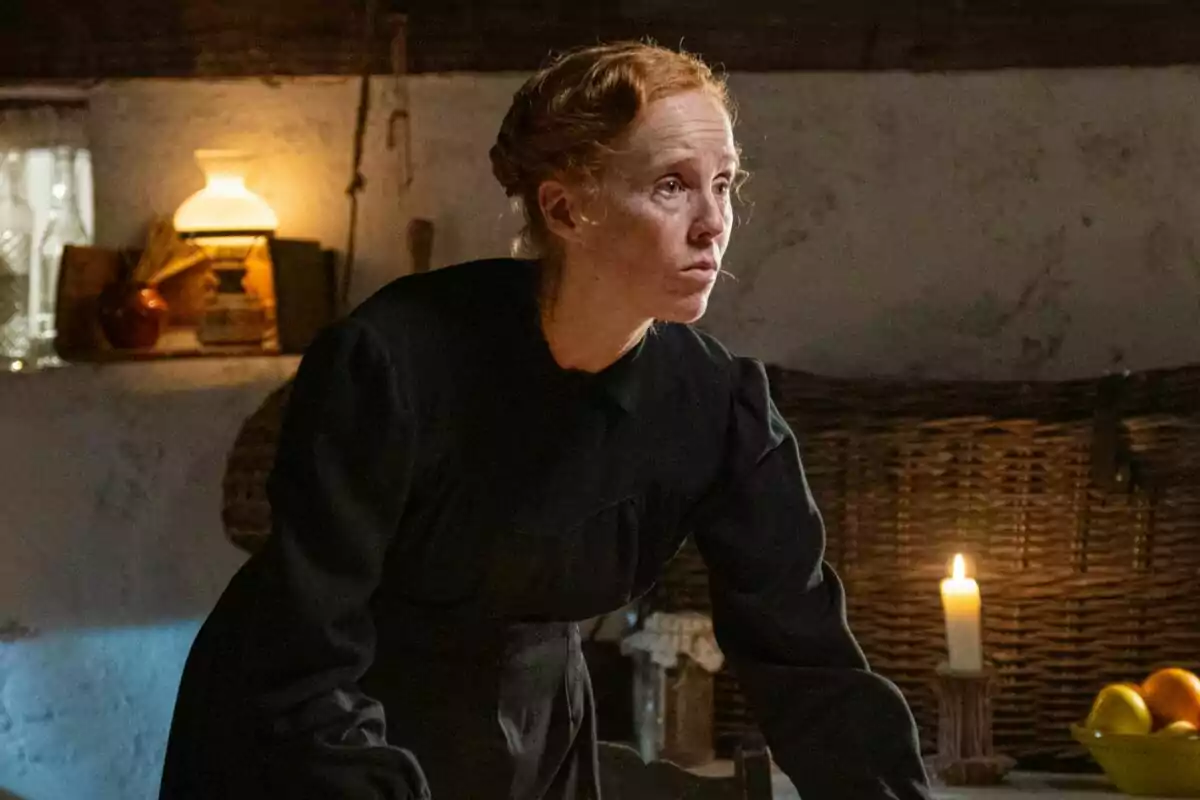 María Castro com a Pla amb cabell recollit i vestit fosc està en una habitació rústica il·luminada per una espelma i un llum, amb una cistella i fruites al fons a La Promesa. María Castro com a Pla amb cabell recollit i vestit fosc està en una habitació rústica il·luminada per una espelma i un llum, amb una cistella i fruites al fons a La Promesa.