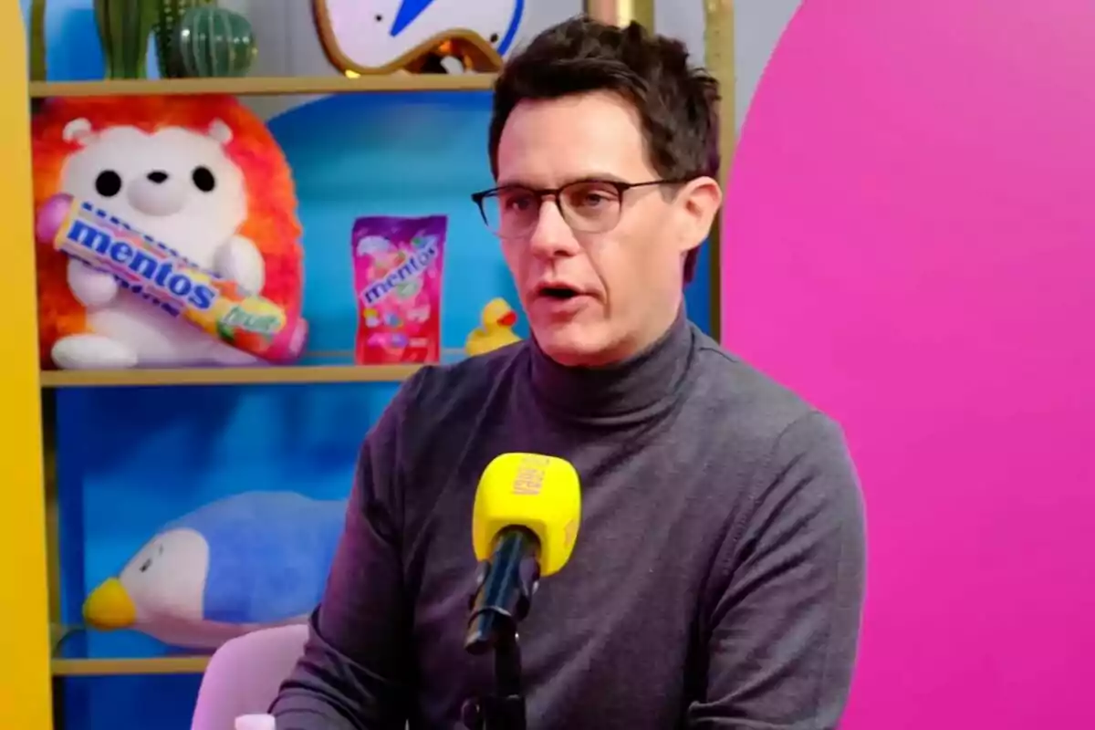 Christian Gálvez con gafas y suéter gris habla frente a un micrófono amarillo en 'Vaya Vaina', un estudio colorido con juguetes y productos en el fondo. Christian Gálvez con gafas y suéter gris habla frente a un micrófono amarillo en 'Vaya Vaina', un estudio colorido con juguetes y productos en el fondo.