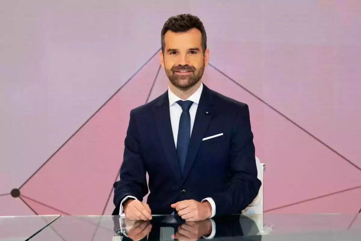 Jon Ariztimuño amb vestit fosc i corbata blava està assegut darrere d'un escriptori de vidre amb un fons geomètric.