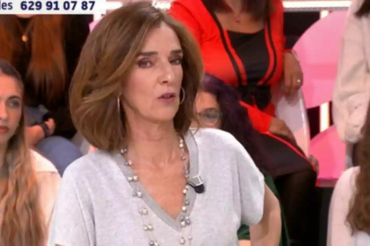 Paloma García-Pelayo amb cabell castany i un micròfon a la roba està en un programa de televisió, amb persones assegudes al fons i un número de telèfon visible a la part superior.