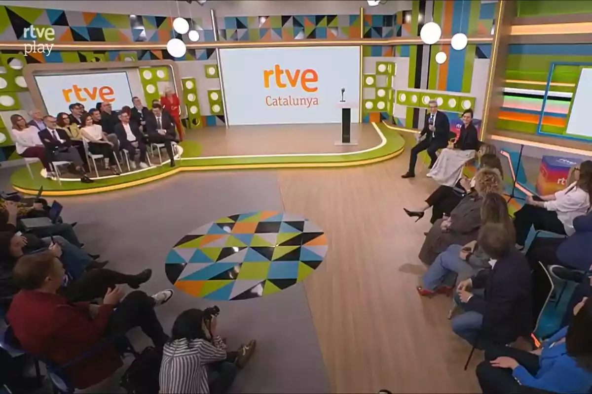 Un grupo de personas está reunido en un estudio de televisión colorido con un escenario que muestra el logo de Un grupo de personas está reunido en un estudio de televisión colorido con un escenario que muestra el logo de