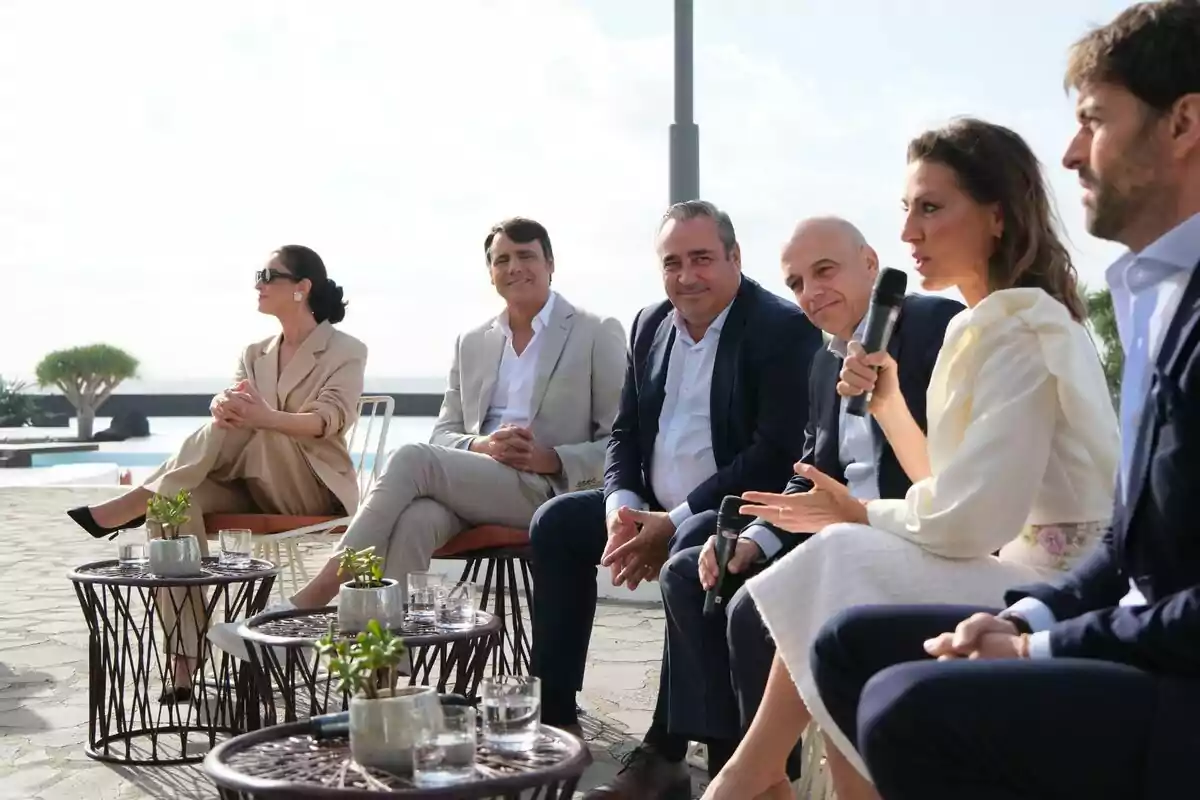 Ion Aramendi, Blanca Romero, Ángeles Blanco y Ricardo Reyes sentadas al aire libre participando en una conversación, con mesas pequeñas y plantas en primer plano. Ion Aramendi, Blanca Romero, Ángeles Blanco y Ricardo Reyes sentadas al aire libre participando en una conversación, con mesas pequeñas y plantas en primer plano.