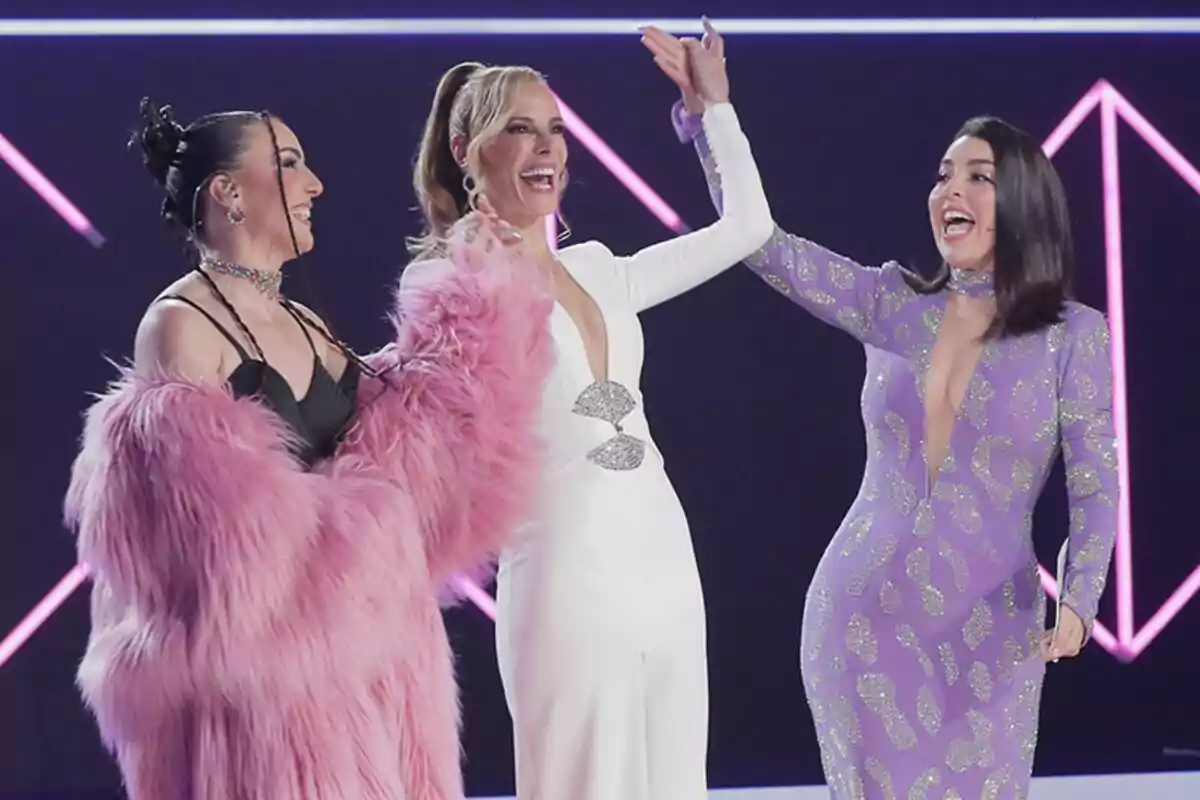 Inés Hernand, Paula Vázquez y Ruth Lorenzo posan juntas en un escenario del Benidorm Fest con luces de neón, una lleva un abrigo rosa peludo, otra un vestido blanco y la tercera un vestido morado con detalles brillantes. Inés Hernand, Paula Vázquez y Ruth Lorenzo posan juntas en un escenario del Benidorm Fest con luces de neón, una lleva un abrigo rosa peludo, otra un vestido blanco y la tercera un vestido morado con detalles brillantes.
