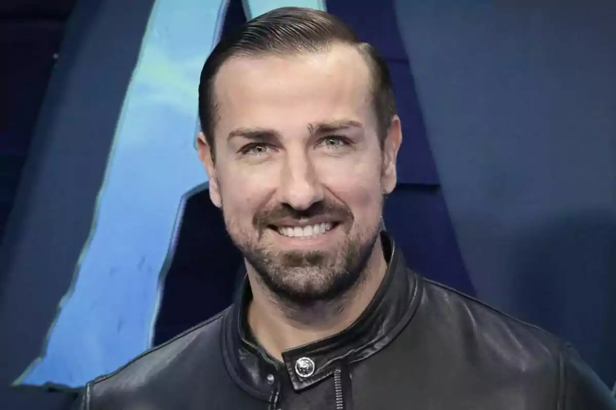 Primer plano de Rafa Mora sonriendo con chaqueta de cuero negra frente a un fondo azul. Primer plano de Rafa Mora sonriendo con chaqueta de cuero negra frente a un fondo azul.