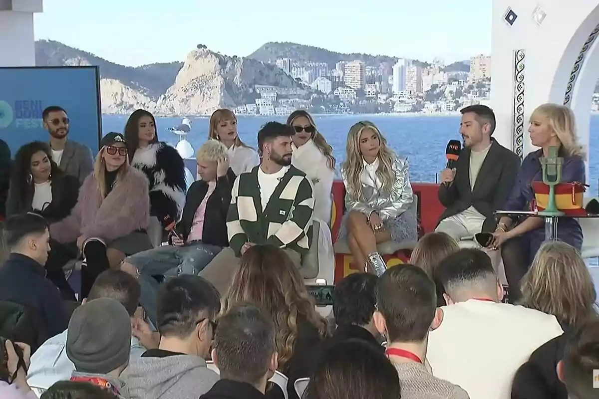 Un grupo de personas está sentado en un escenario frente a un público, con un fondo de mar y montañas, durante la rueda de prensa del Benidorm Fest.