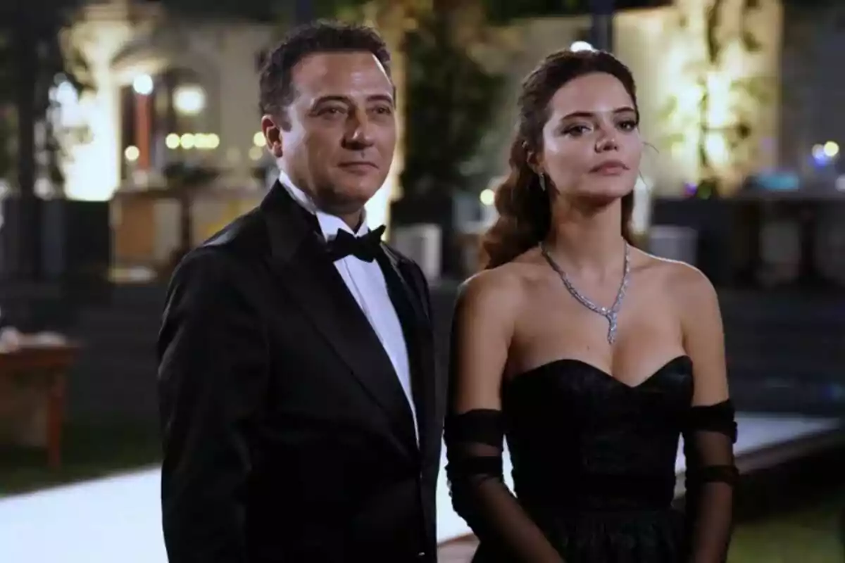 Fotografía de una pareja en Una vida perfecta elegantemente vestida, Yiğit Özşener con un esmoquin negro y Hilal Altınbilek con un vestido negro sin tirantes y un collar de diamantes, posando en un evento nocturno. Fotografía de una pareja en Una vida perfecta elegantemente vestida, Yiğit Özşener con un esmoquin negro y Hilal Altınbilek con un vestido negro sin tirantes y un collar de diamantes, posando en un evento nocturno.