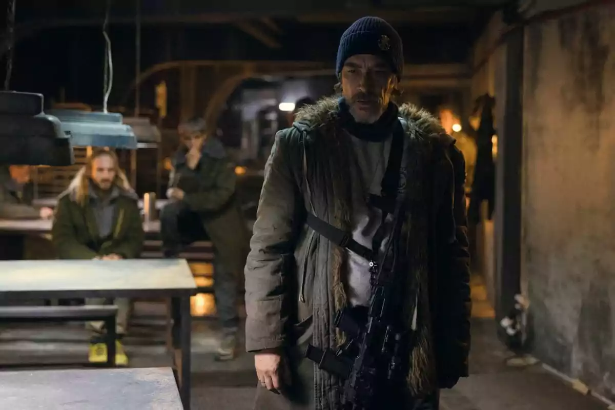 Óscar Jaenada con gorro y abrigo oscuro sostiene un arma en una habitación con poca luz, mientras otras dos personas están sentadas al fondo en Punto Nemo.