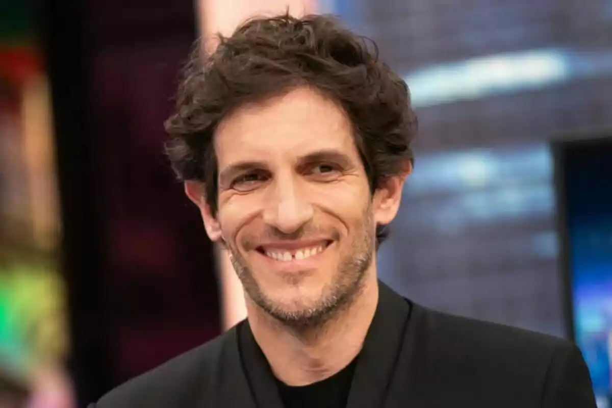 Quim Gutiérrez con cabello rizado y barba ligera sonríe mientras lleva una chaqueta negra, con un fondo desenfocado de colores en 'El Hormiguero'.
