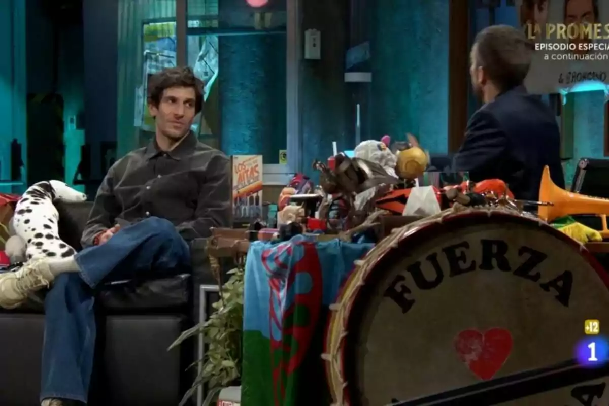 Quim Gutiérrez está sentado en un sofá en La Revuelta junto a David Broncano de espaldas, rodeado de varios objetos coloridos y un tambor grande con la palabra Quim Gutiérrez está sentado en un sofá en La Revuelta junto a David Broncano de espaldas, rodeado de varios objetos coloridos y un tambor grande con la palabra