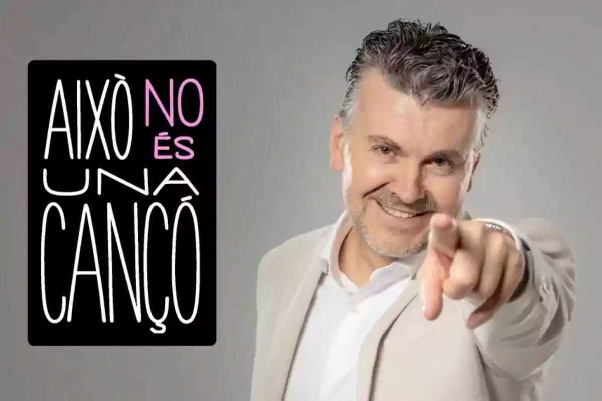 Ramon Gener somrient amb barba i cabell canós, vestit amb un vestit clar, assenyala cap a la cambra; al seu costat hi ha un cartell negre amb lletres blanques i roses que diu “Això no és una cançó”.