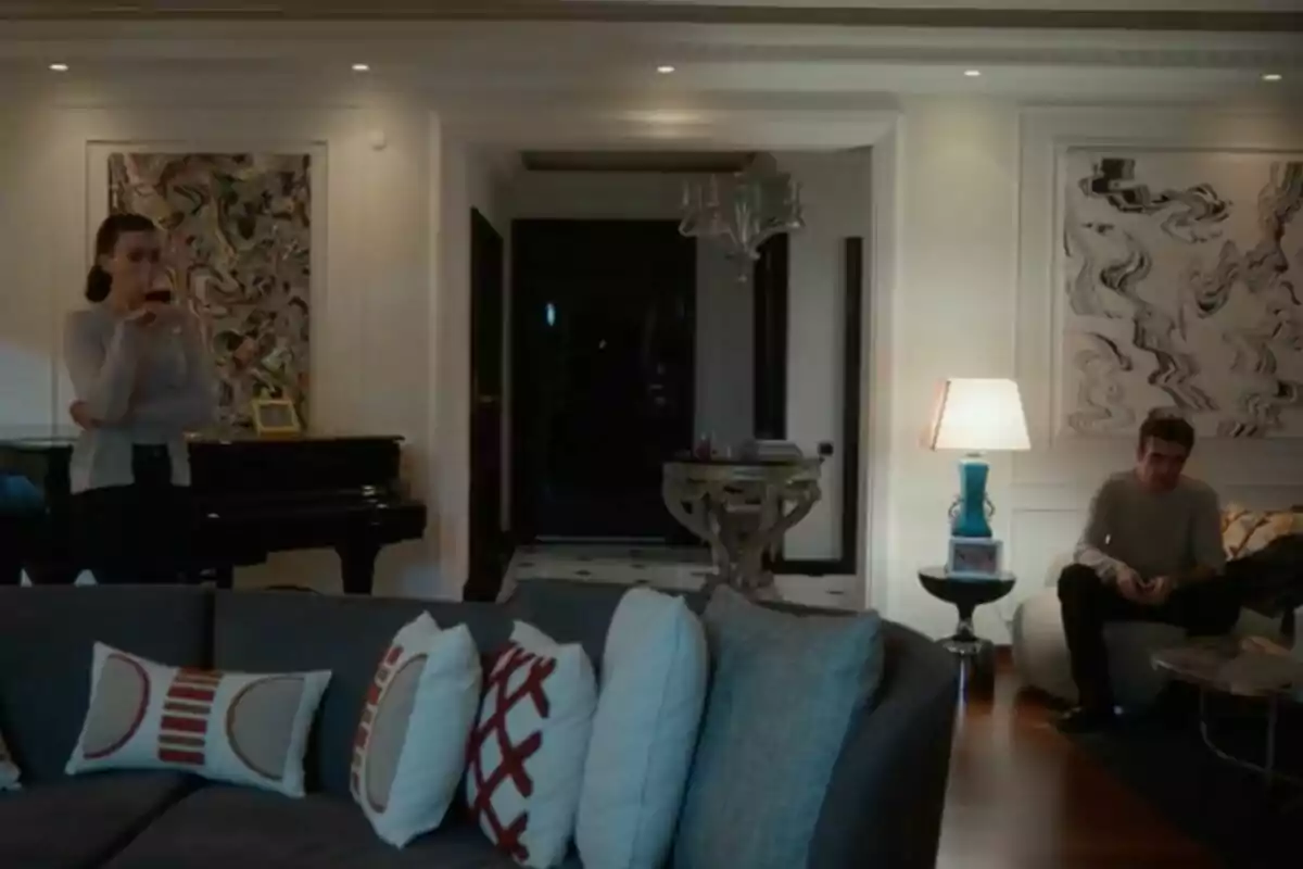 Una sala d'estar moderna amb un sofà gris i coixins decoratius, una dona dreta al costat d'un piano i un home assegut a prop d'una làmpada, amb art abstracte a les parets a la sèrie 'Renaixement'. Una sala d'estar moderna amb un sofà gris i coixins decoratius, una dona dreta al costat d'un piano i un home assegut a prop d'una làmpada, amb art abstracte a les parets a la sèrie 'Renaixement'.