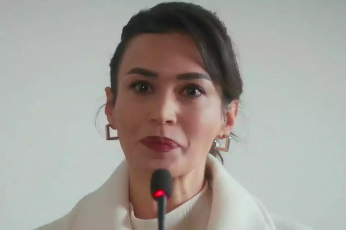 Rengin de cabello oscuro y labios rojos hablando frente a un micrófono con fondo claro en Renacer