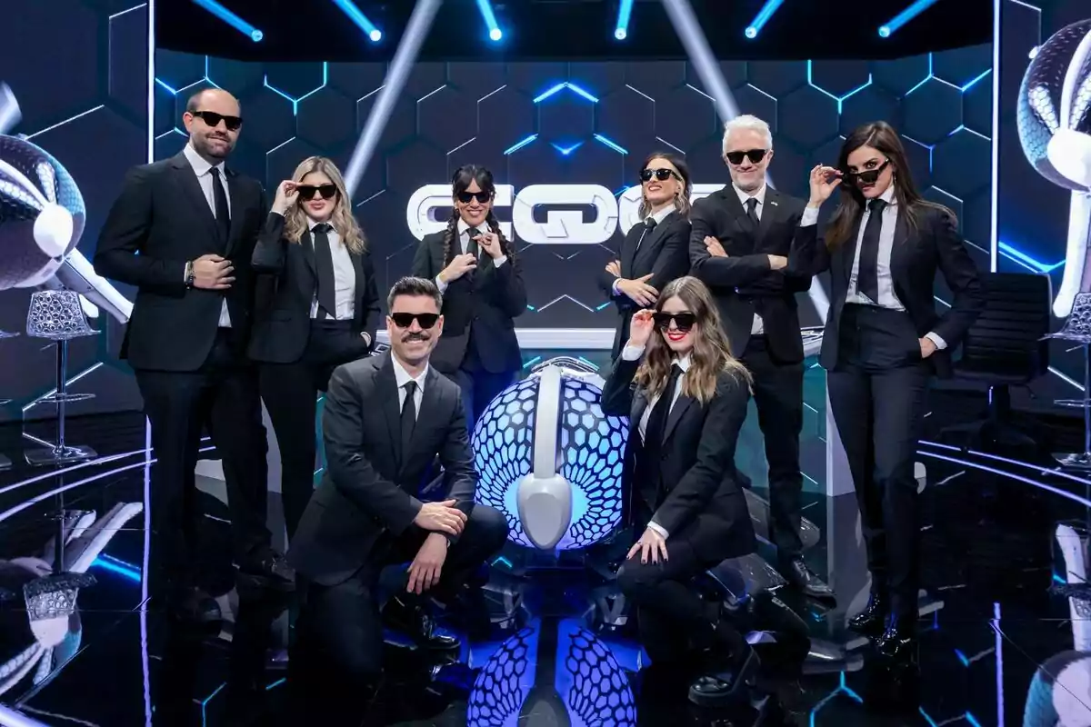 Paula Púa, Dani Fez, Violeta Muñoz, Ana Francisco, Luis Fabra, Irene Junquera, Alba Moreno y Andy K vestidos con trajes negros y gafas de sol posan en el plató de Caiga quien caiga futurista con luces azules y un fondo geométrico. Paula Púa, Dani Fez, Violeta Muñoz, Ana Francisco, Luis Fabra, Irene Junquera, Alba Moreno y Andy K vestidos con trajes negros y gafas de sol posan en el plató de Caiga quien caiga futurista con luces azules y un fondo geométrico.