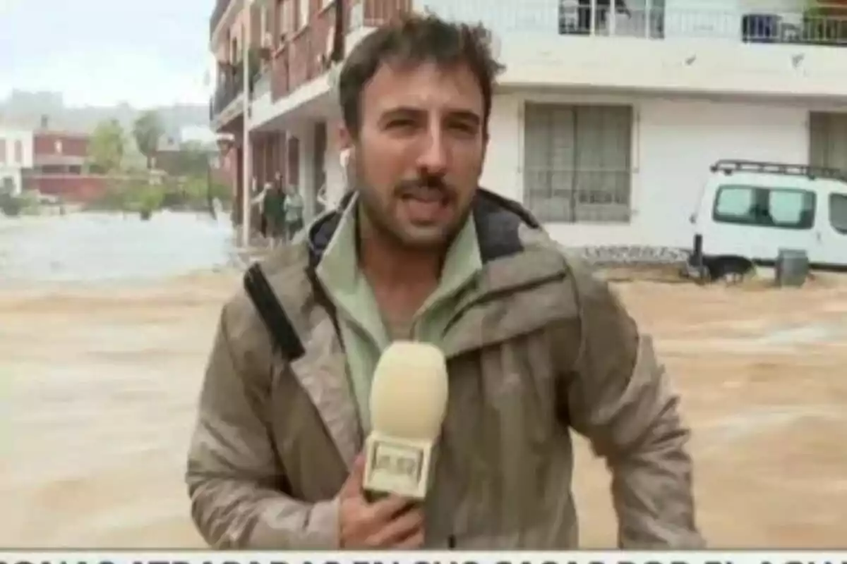 Un reporter de TardeAR amb jaqueta impermeable sosté un micròfon mentre informa des d?una zona inundada amb edificis i un vehicle al fons. Un reporter de TardeAR amb jaqueta impermeable sosté un micròfon mentre informa des d?una zona inundada amb edificis i un vehicle al fons.